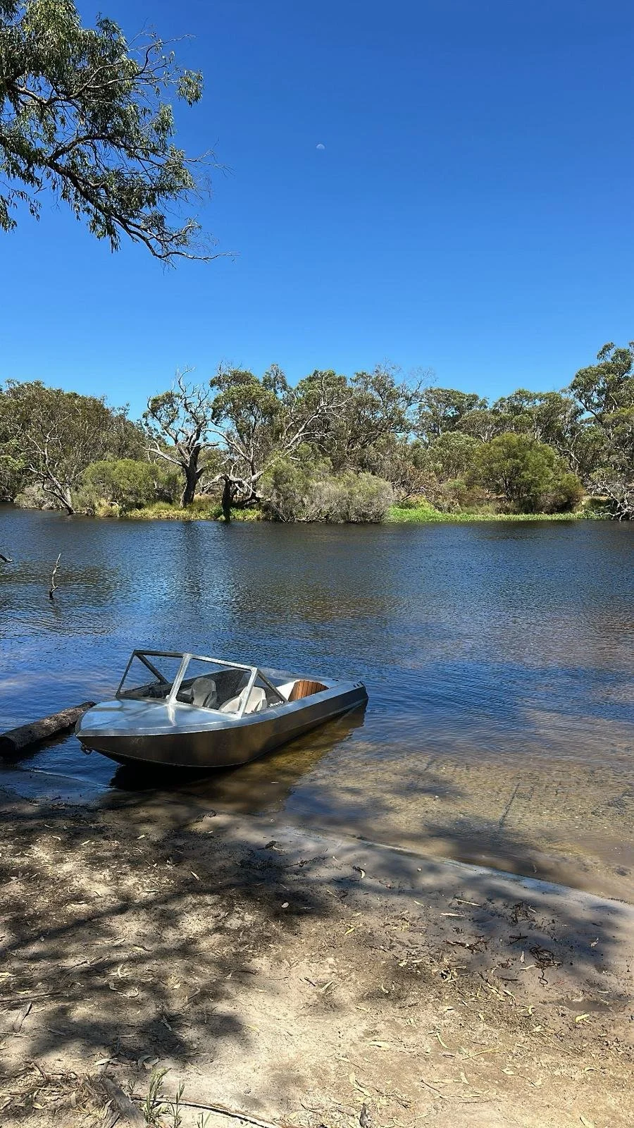 A small boat floating in a river with trees on the opposite bank and a clear blue sky above, featuring a faint moon.