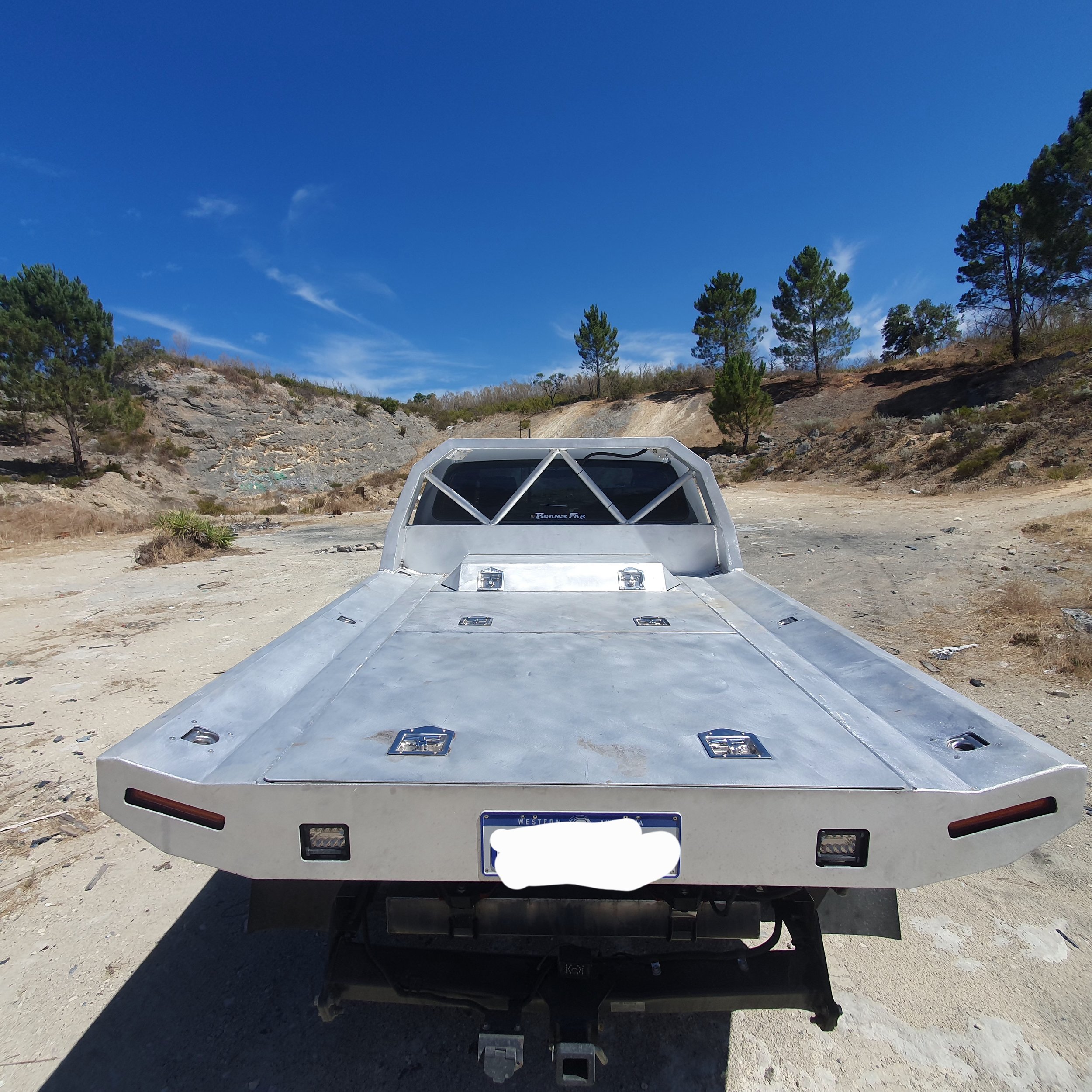 A metallic vehicle with a flatbed and a front windscreen, parked on a dirt area with trees and rocky hills in the background under a blue sky.