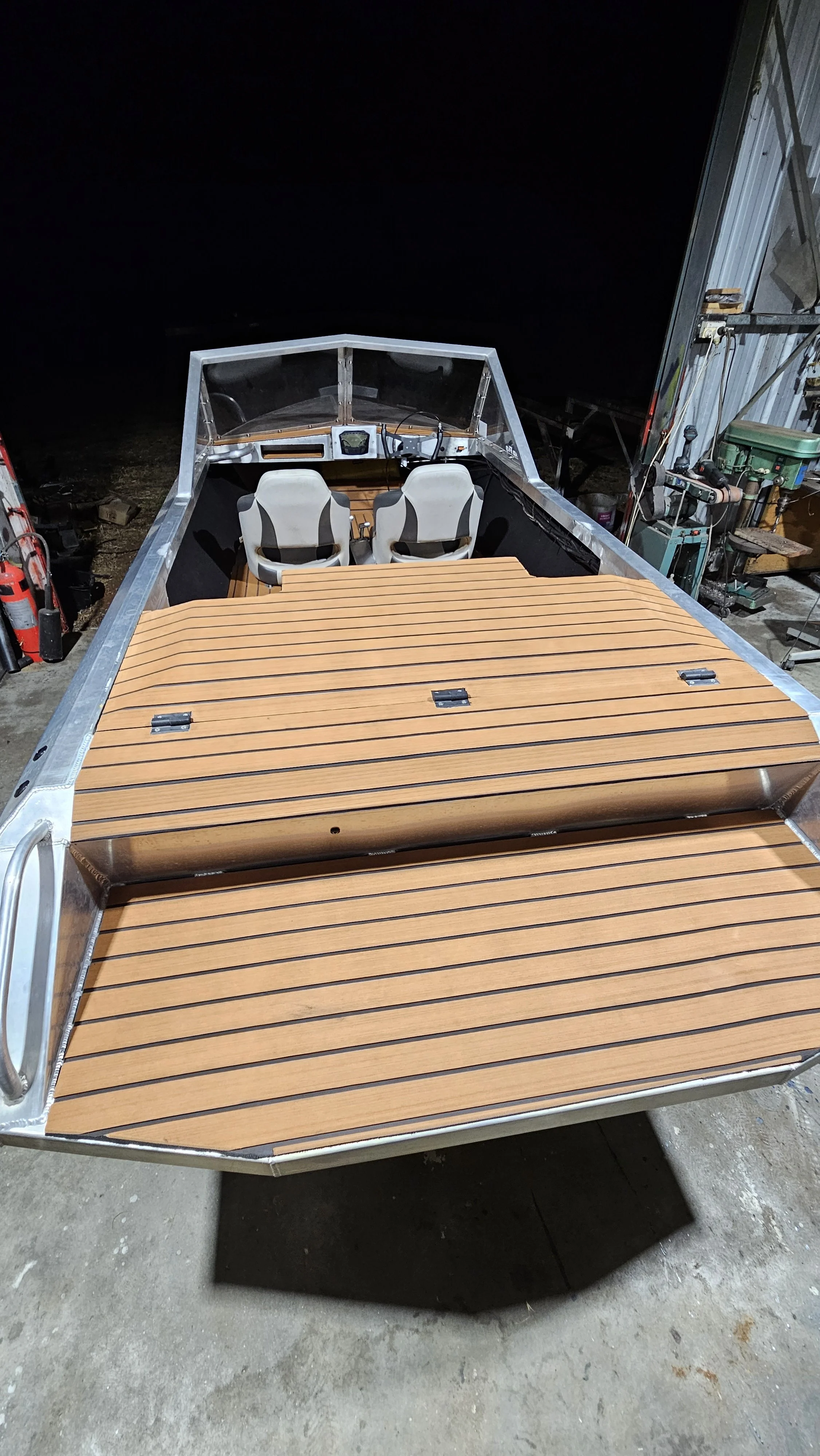 Front view of a boat with a wooden deck and two steering chairs, parked inside a garage at night.