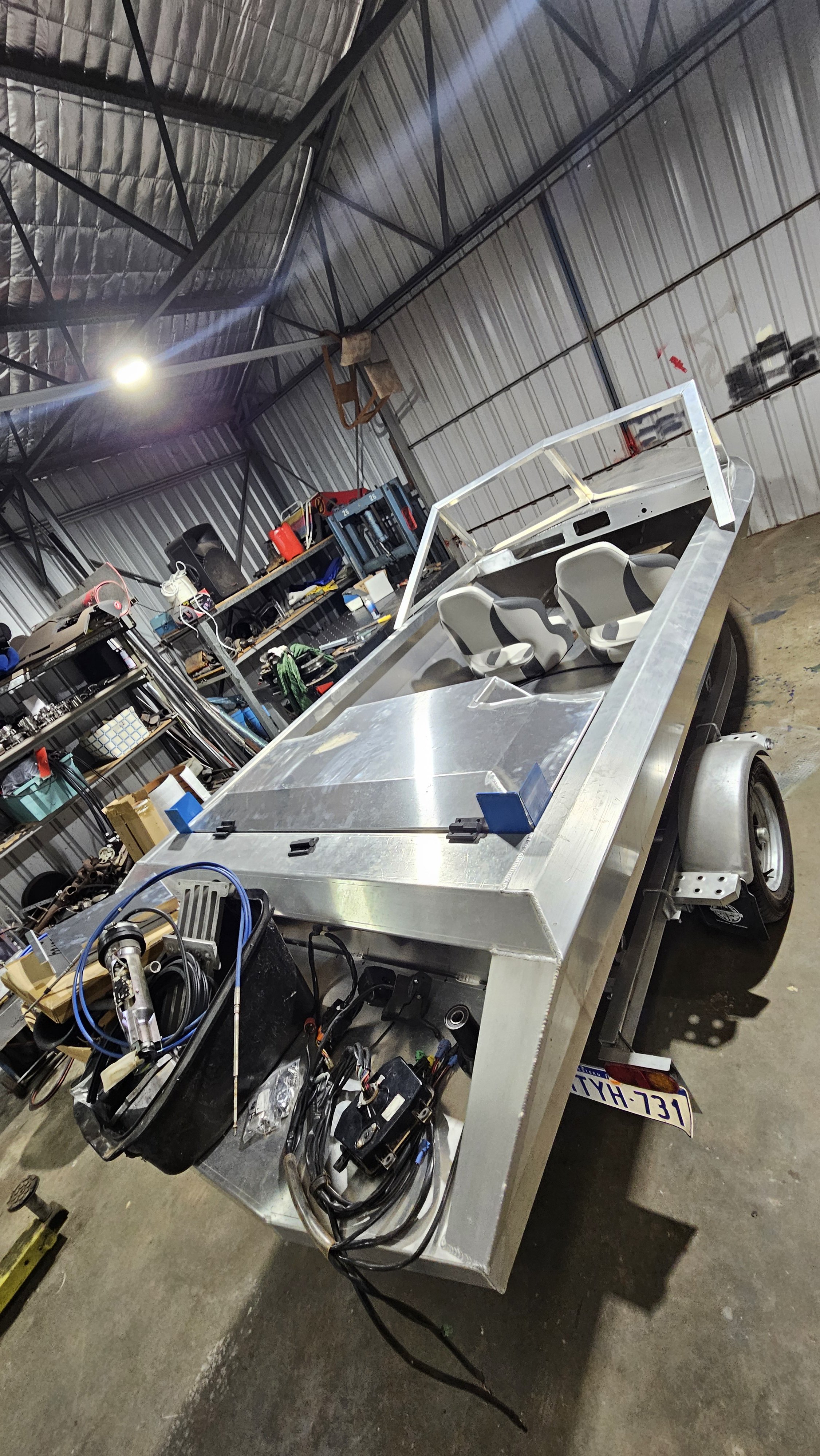 An unfinished aluminum vehicle frame in a workshop with various tools and parts on shelves in the background.