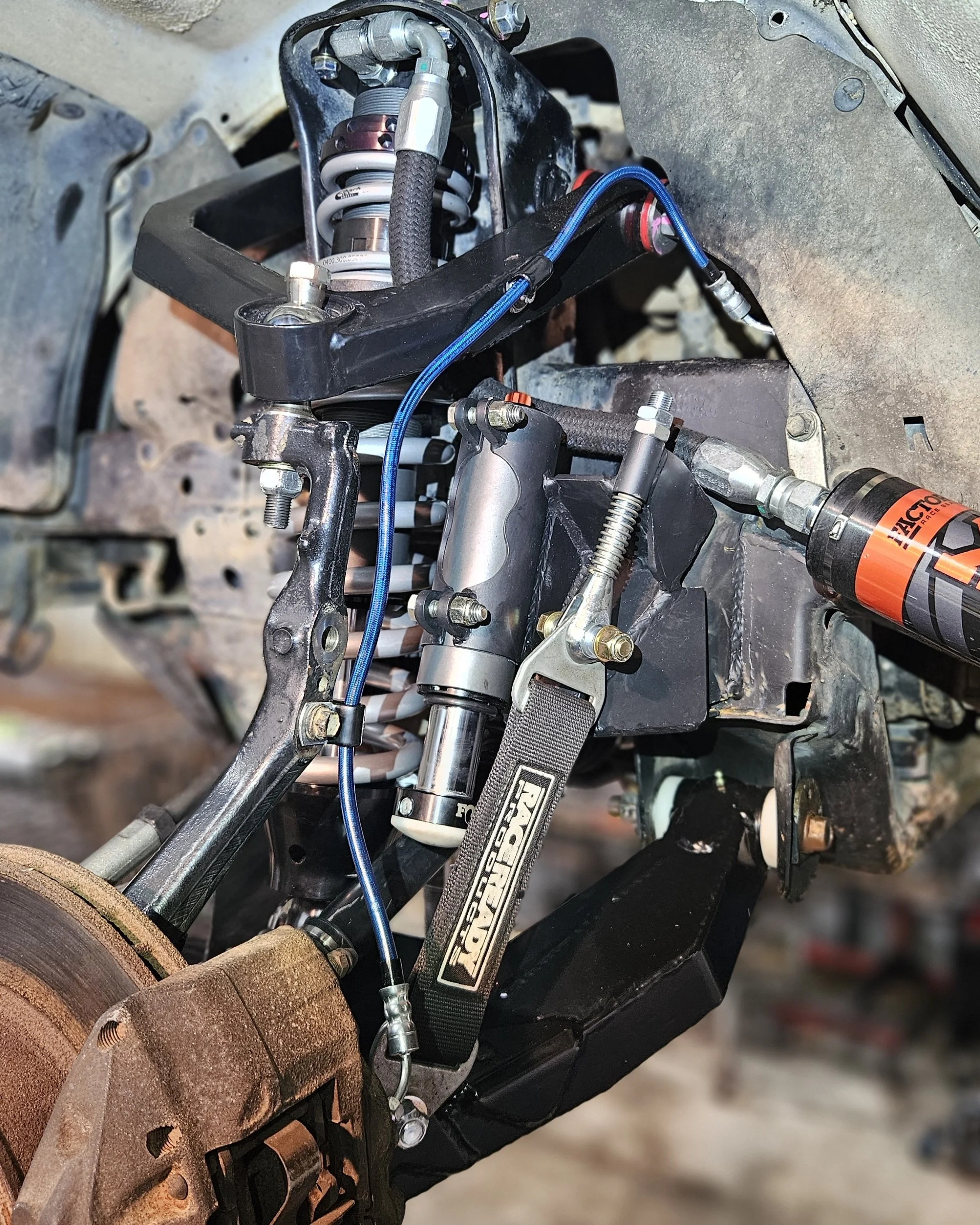 Close-up of a vehicle's suspension and steering components, including shock absorber, control arms, and hydraulic lines, with a rusty brake rotor in the foreground.