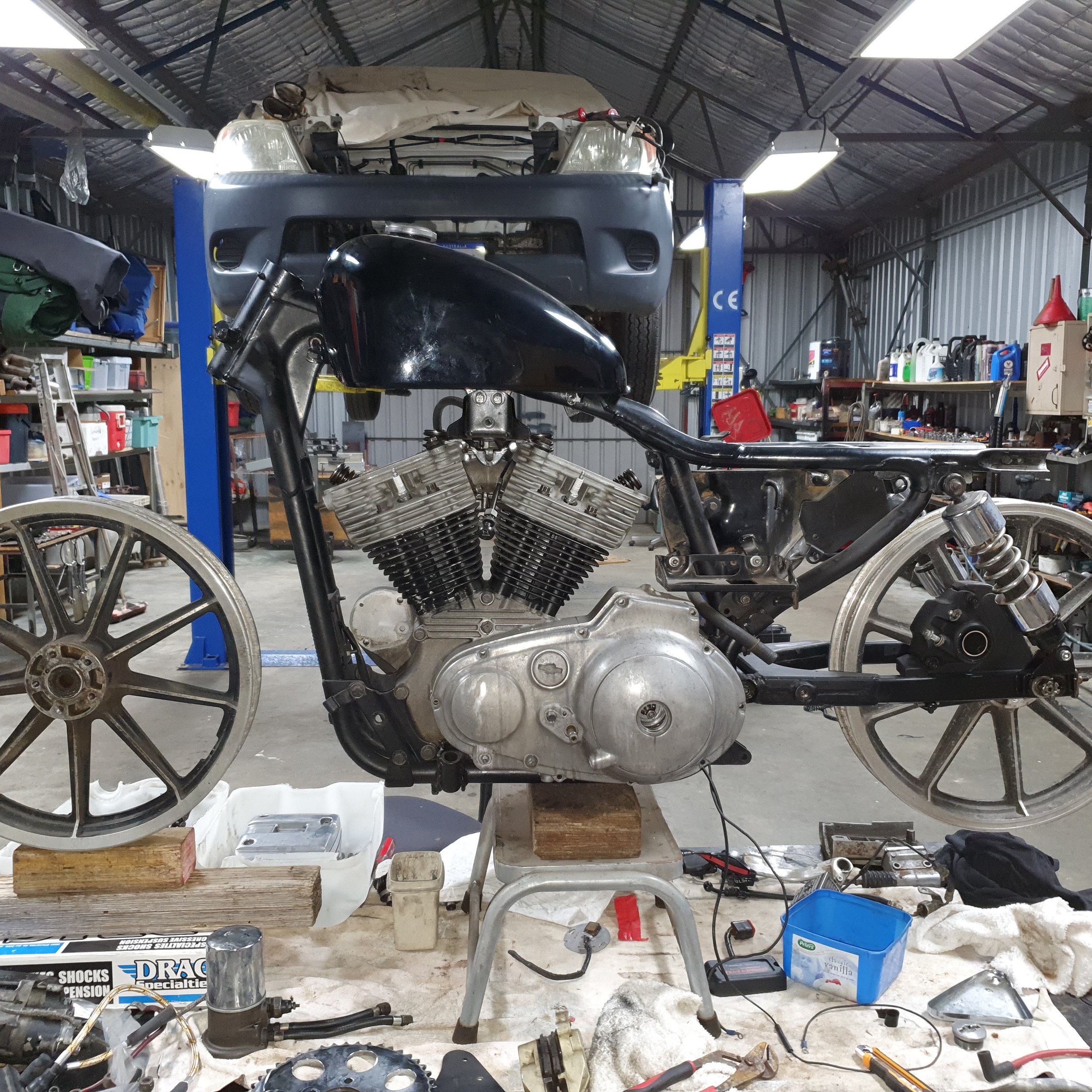 Motorcycle frame with engine installed and front wheel mounted, in a workshop with tools and spare parts around.
