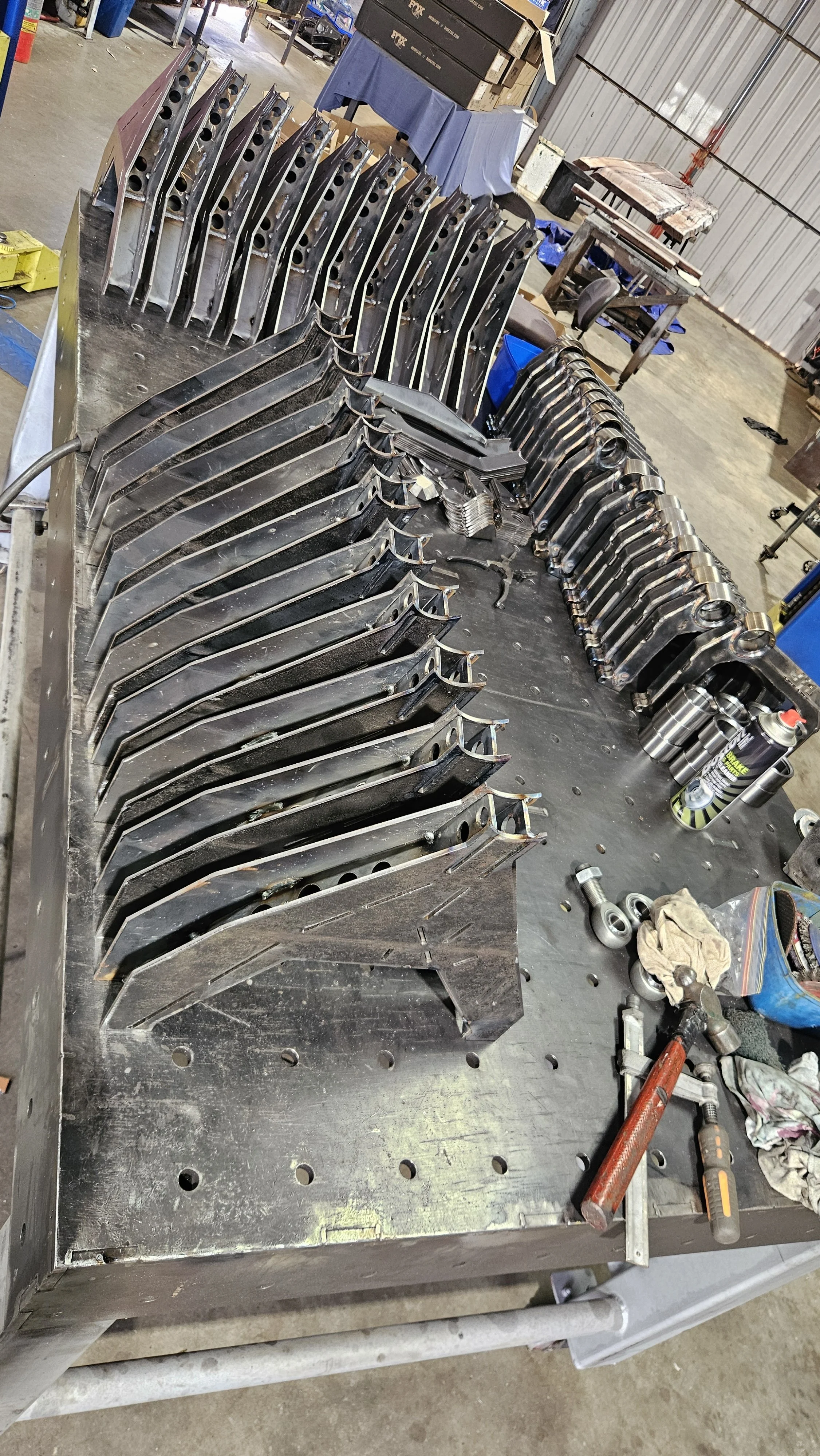 Metal workshop table with multiple curved metal parts, tools, and spray cans, in an industrial setting.