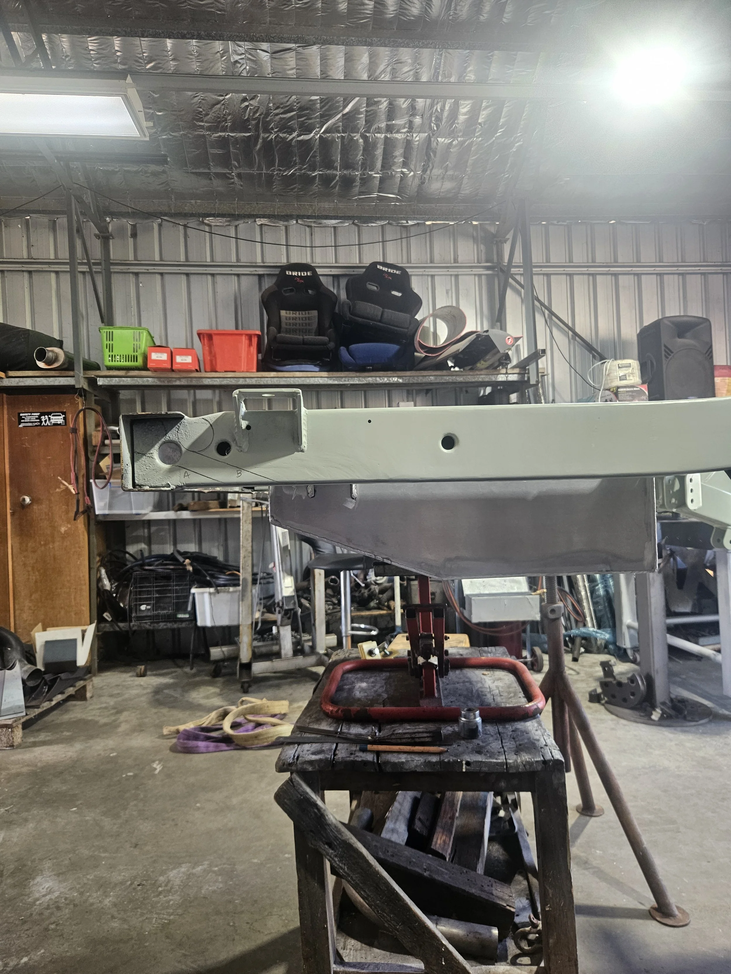 Workshop with a metal frame structure, workbench, and tools. Two racing seats on a shelf, various boxes and equipment around.