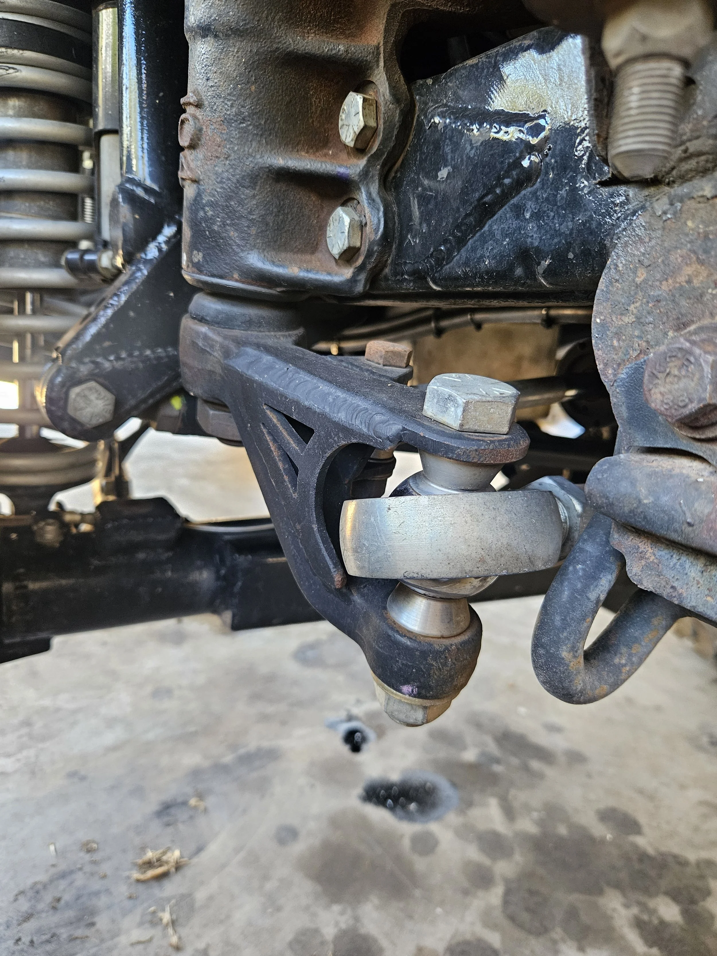 Close-up of a mechanical part on a vehicle, featuring metal bolts, nuts, and a ball joint, with some rust and signs of wear.