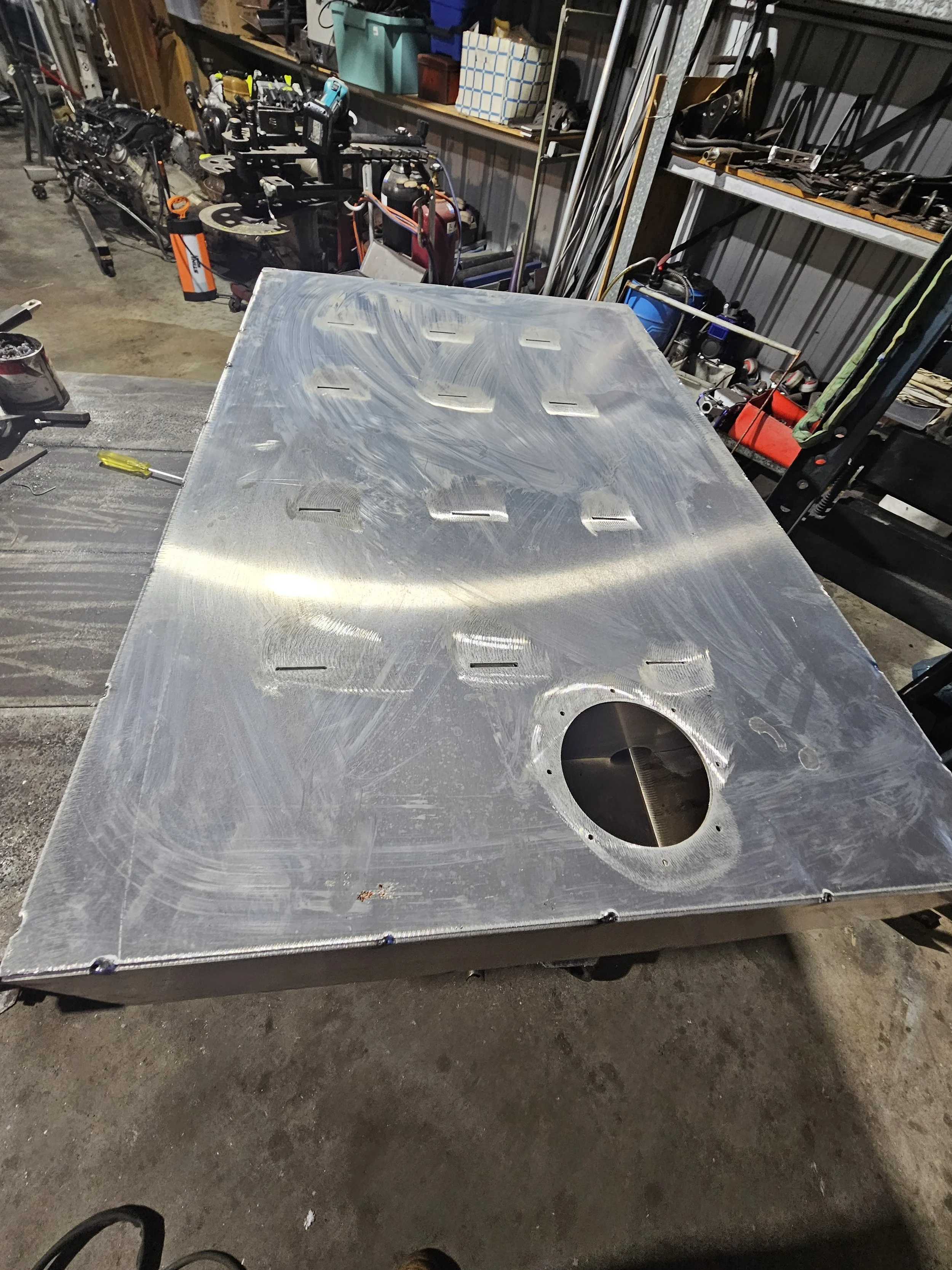 Metal worktable with a large circular hole and a series of evenly spaced smaller rectangular slots, set in a workshop surrounded by tools and equipment.