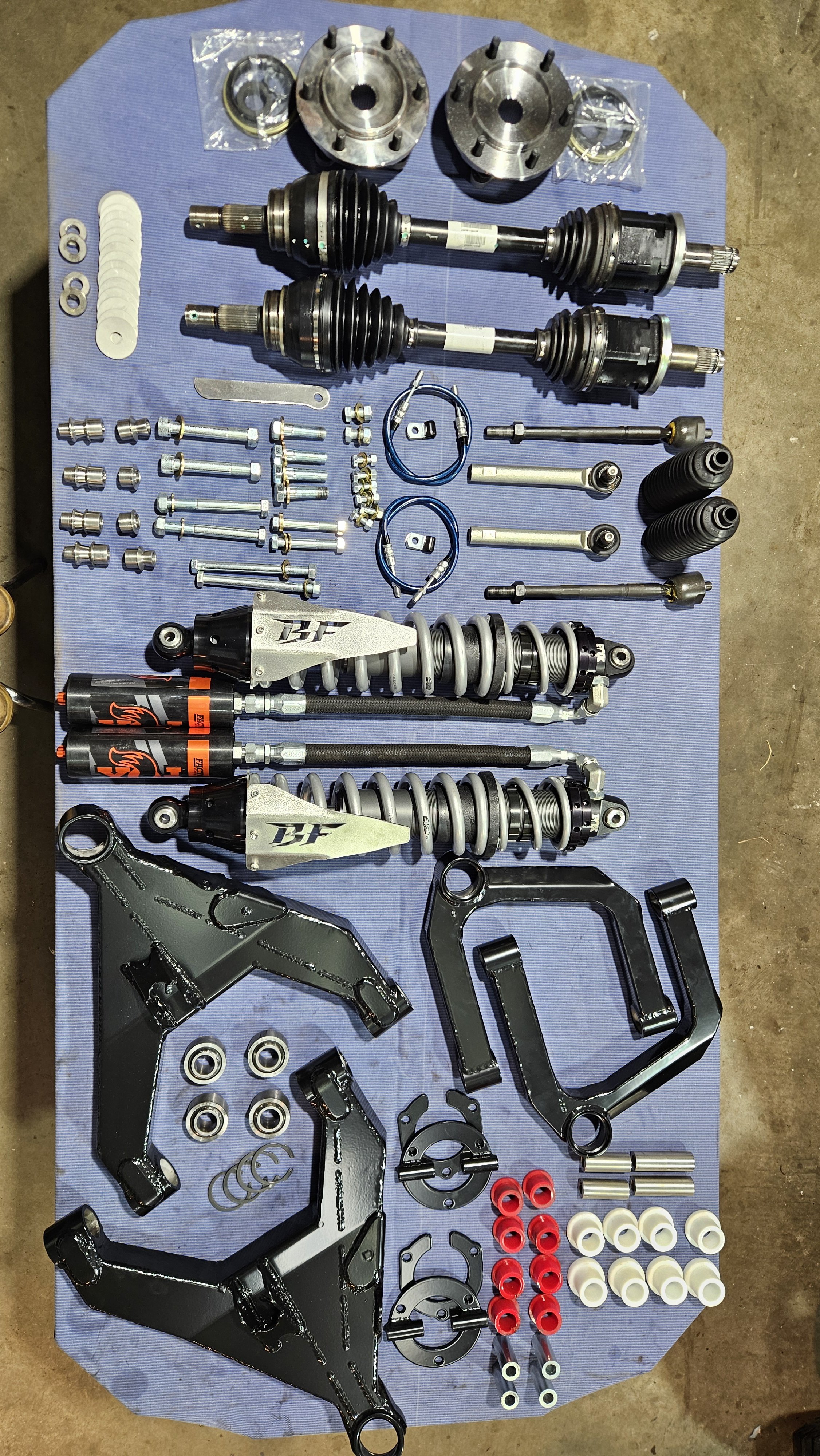 Disassembled vehicle suspension and drivetrain parts laid out on a blue mat, including shock absorbers, control arms, axles, and various bolts and brackets.