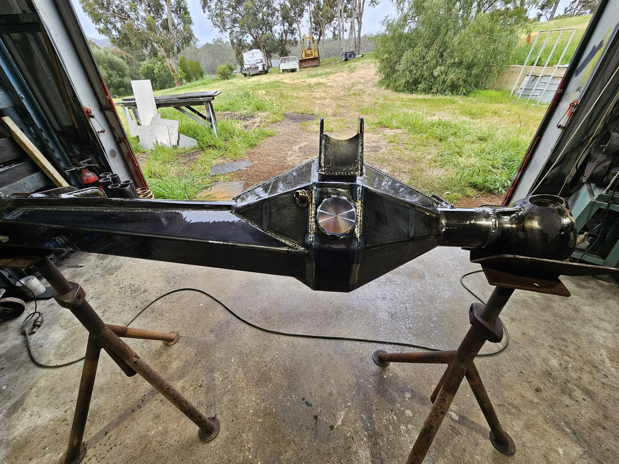 A black metal boat propeller shaft housed on a metal stand inside a garage or workshop, with an outdoor grassy yard and trees visible through the open garage door.