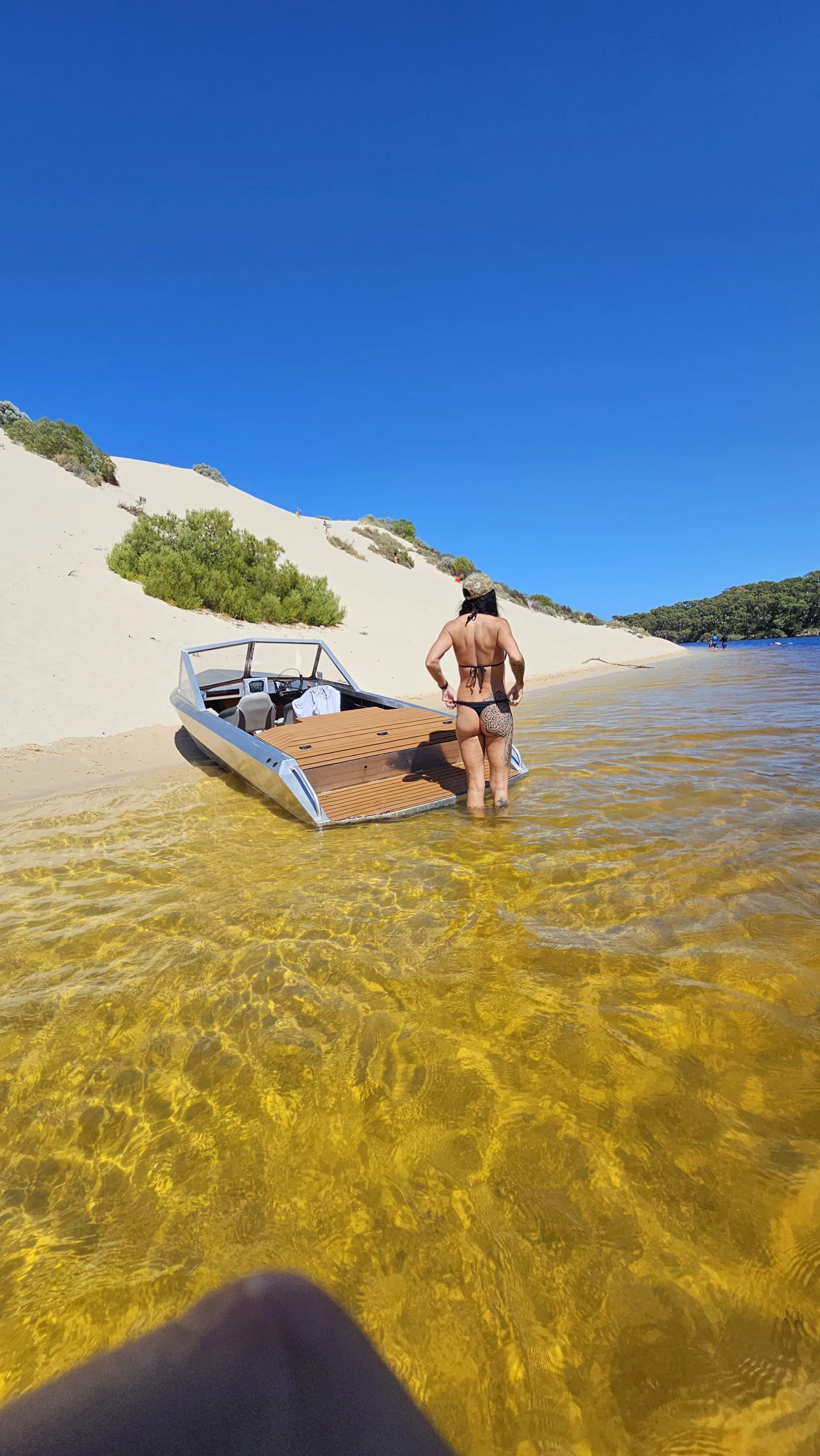 A woman in a bikini standing in clear yellow water next to a small boat on a sandy beach with green bushes and a blue sky.