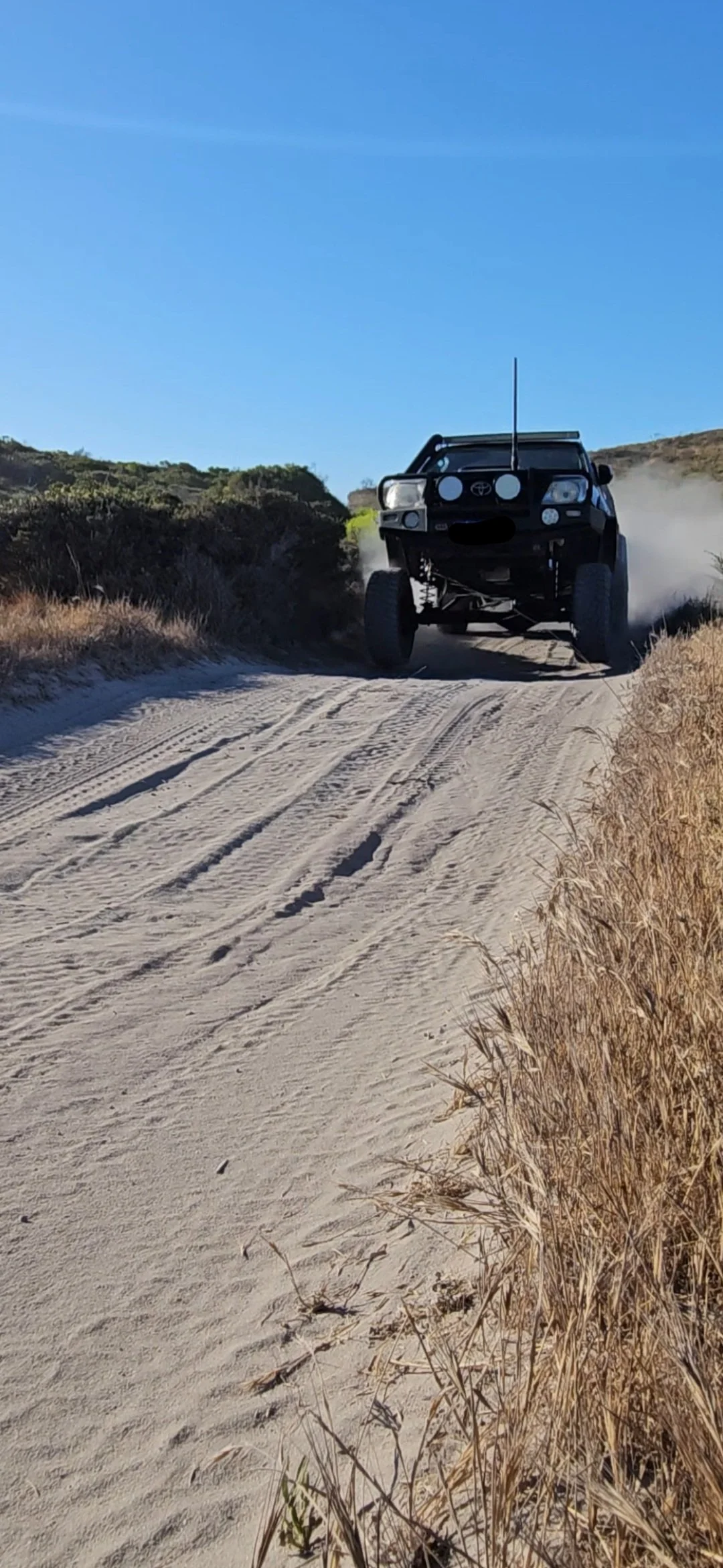 Off-road vehicle driving on a sandy trail with dry grass on the side under a clear blue sky.