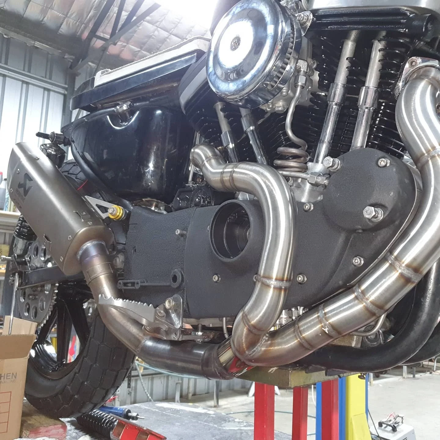 Close-up view of a motorcycle engine being assembled or repaired in a workshop. The engine has shiny metallic parts and a black engine casing. There is a large exhaust pipe and part of the motorcycle's frame is visible, including a rear wheel and a r