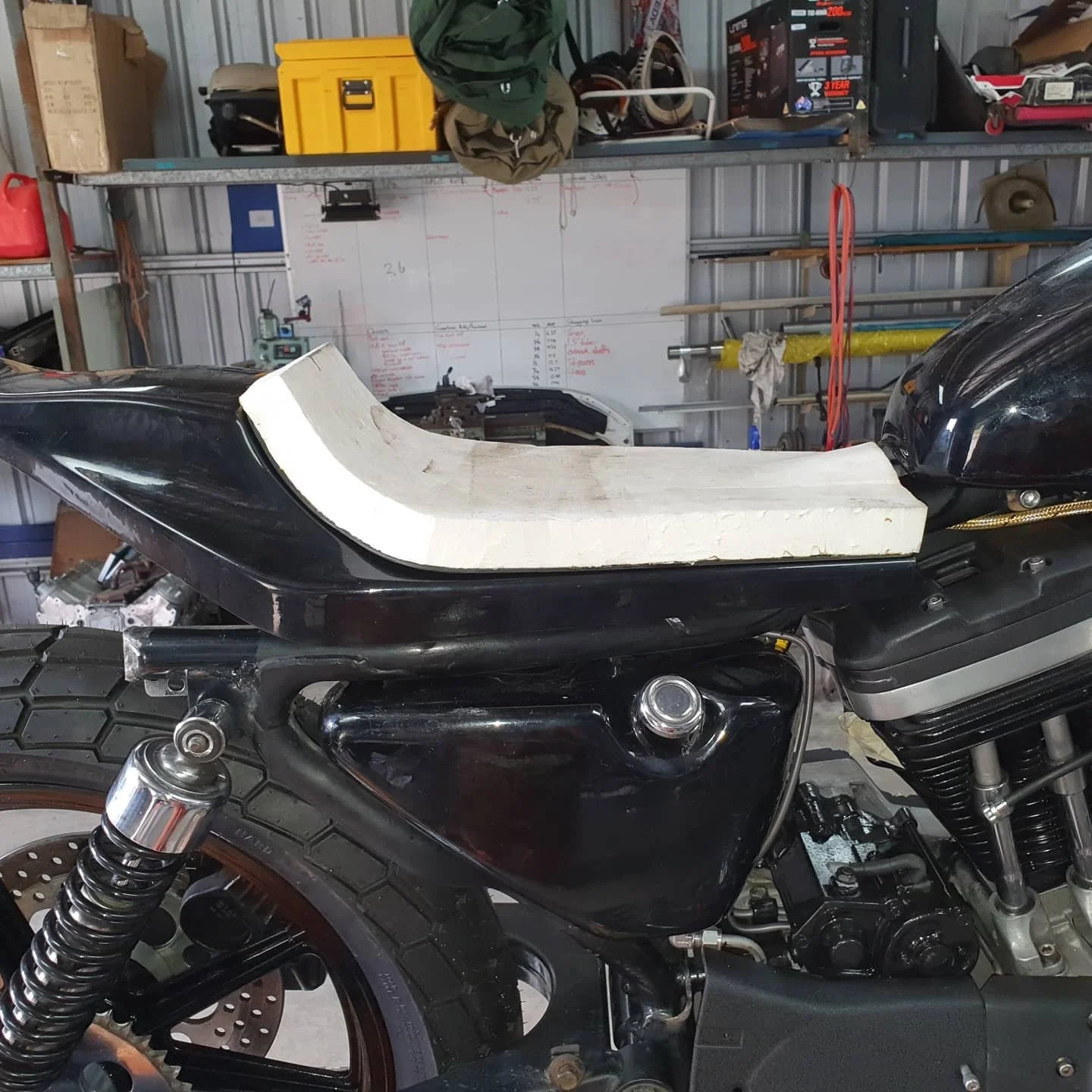 A motorcycle with a white foam or wood piece on top, inside a garage with tools and equipment on the shelves and a whiteboard in the background.
