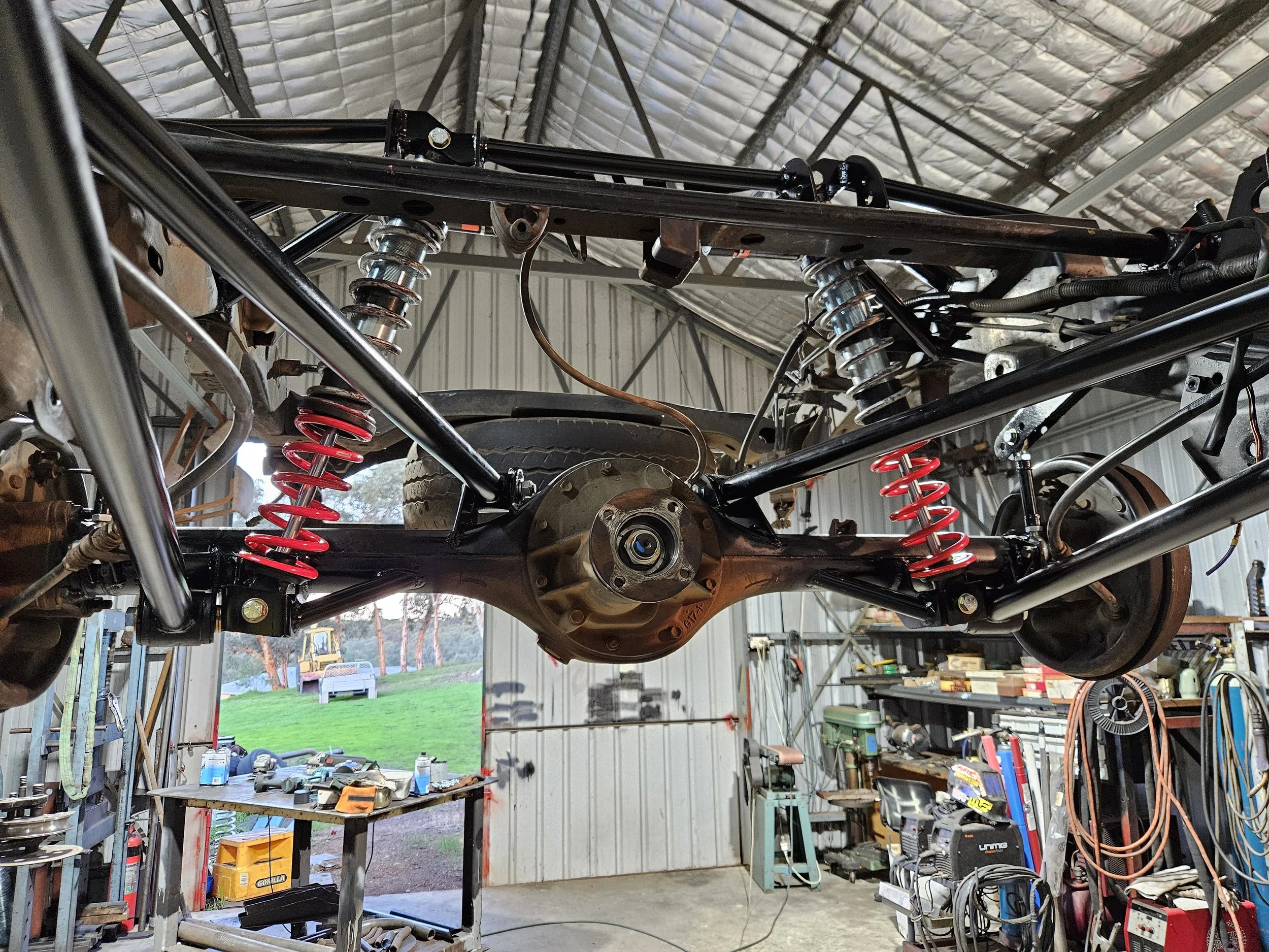 Interior of a garage with a vintage car chassis being assembled or repaired, showing suspension components, coil springs, and other mechanical parts, with workbenches and tools in the background.