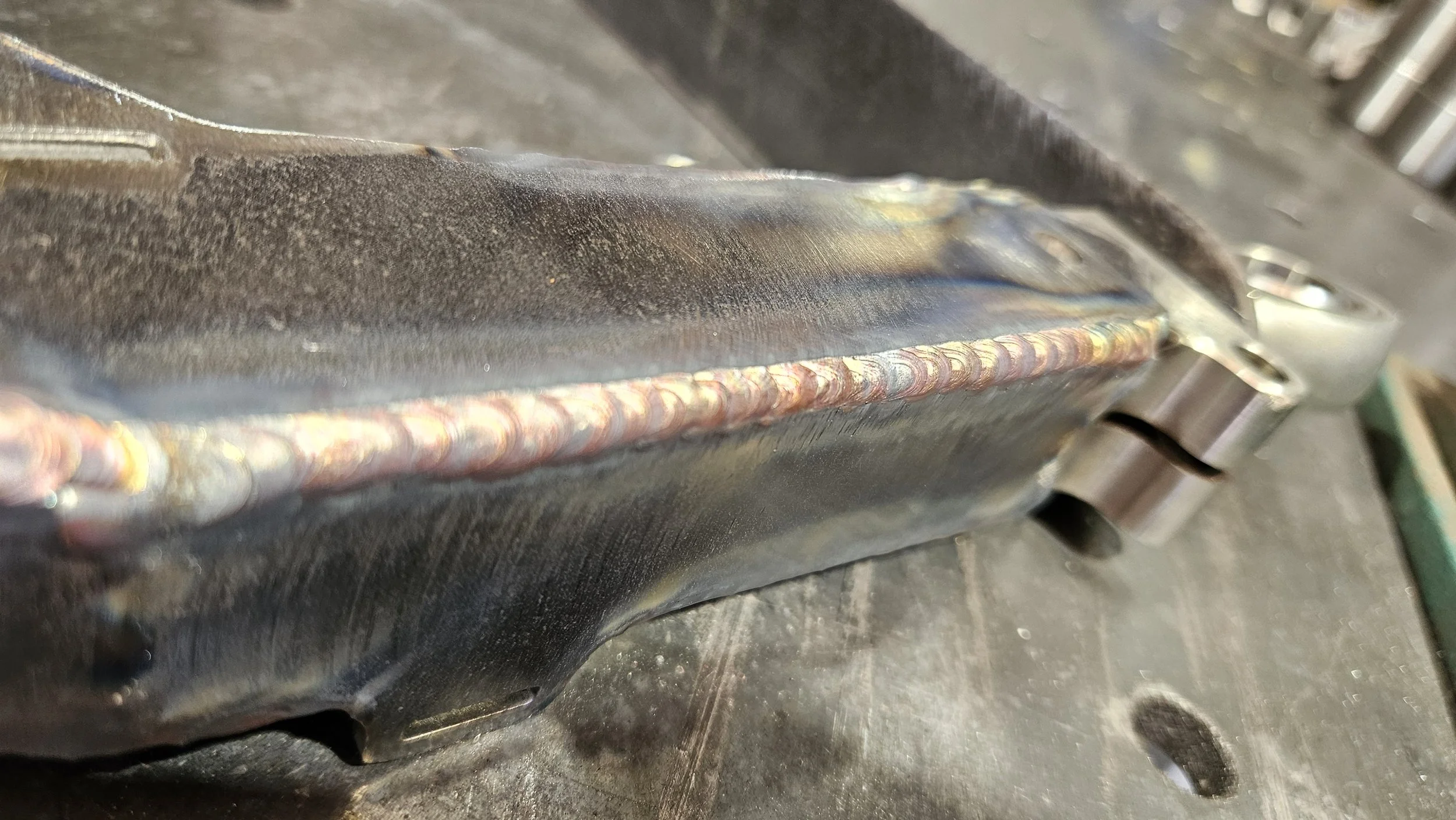 Close-up of a metal component with visible welding bead along the seam, held in a workshop environment with a workbench in the background.