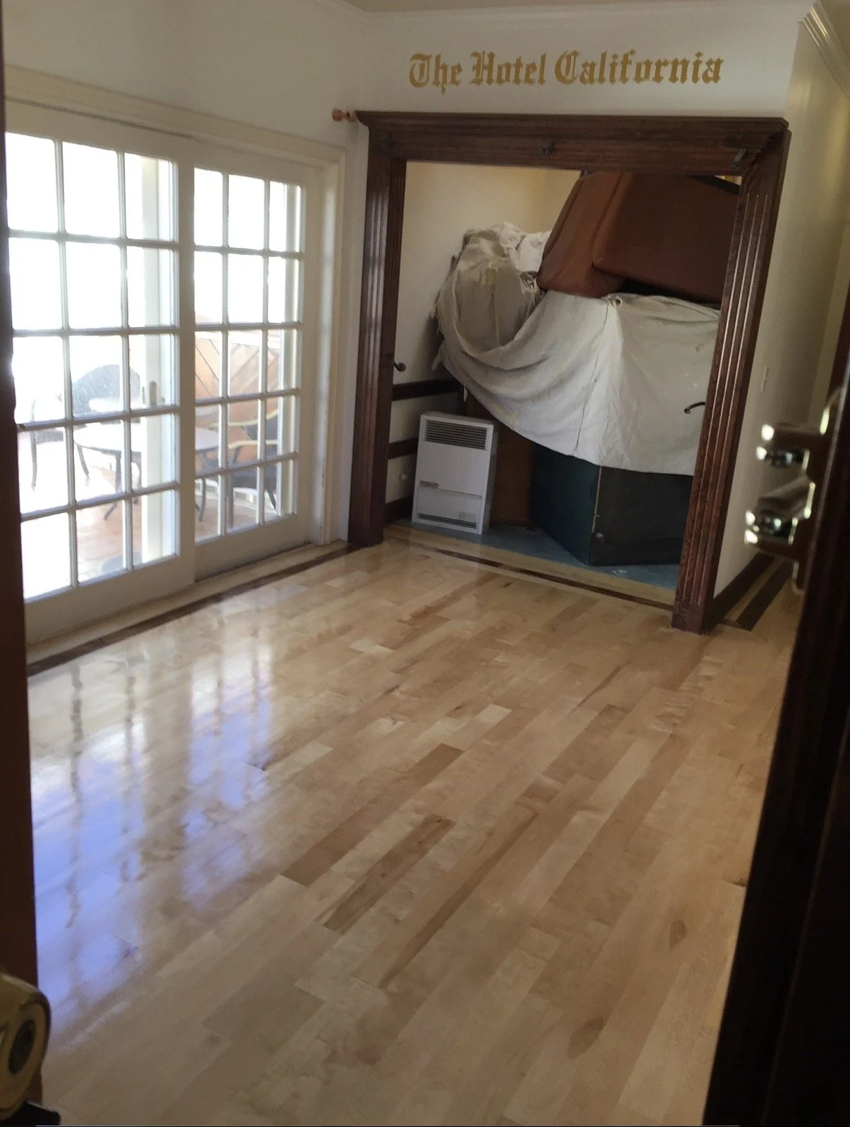 A small room with wooden flooring, glass-paneled doors leading to an outdoor area, and a built-in nook with a bed or furniture covered in a white sheet. The room has a sign above the nook that reads 'The Hotel California' in gold lettering.