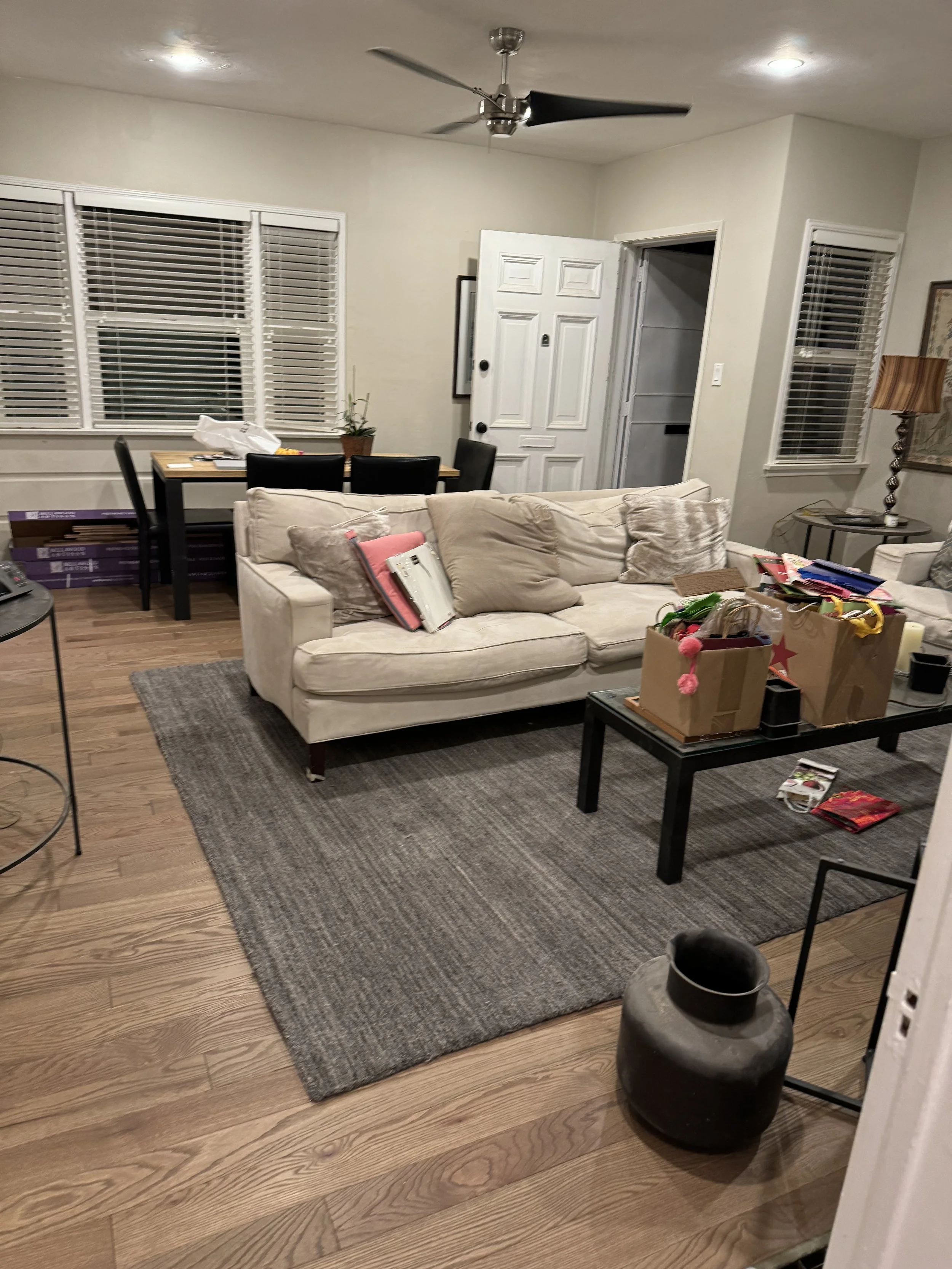 Living room with beige sofa, gray area rug, black coffee table with boxes and various items, dining area with black chairs and wooden table, large windows with white blinds, wooden flooring, ceiling fan, and a door open to a pantry.