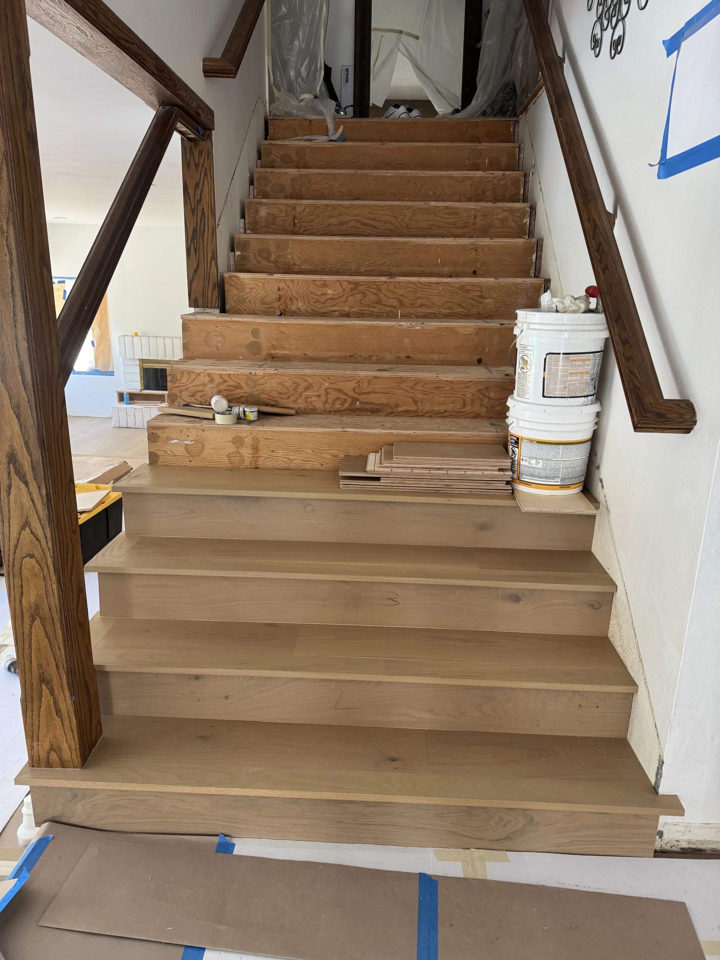 Interior view of a staircase under renovation with natural wood stairs, some painted or treated and others still raw, construction materials including paint buckets, wood planks, tools, and masking tape around the area.