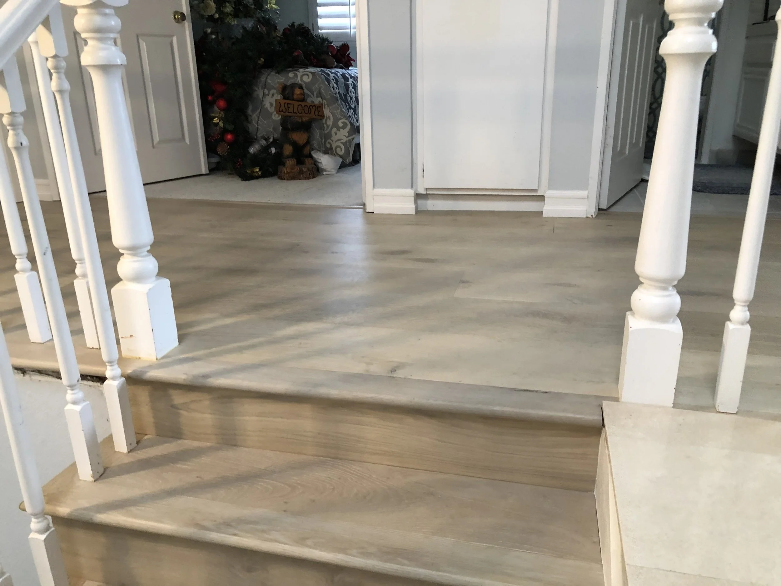 Inside view of a staircase with wooden steps and white balusters, leading to a room with Christmas decorations and a Christmas tree visible in the background.