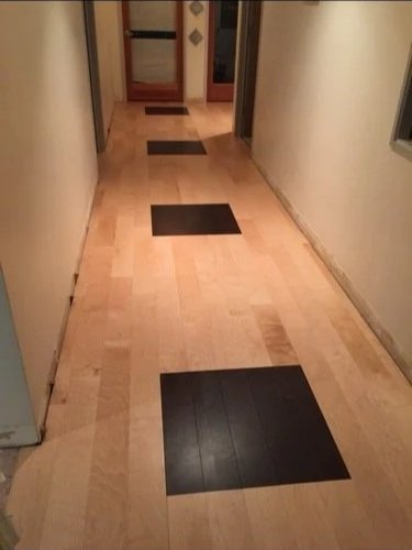 A narrow hallway with light-colored wooden flooring and three rectangular black rugs. The hallway has beige walls and leads to a door at the end with glass panels and a wooden frame.