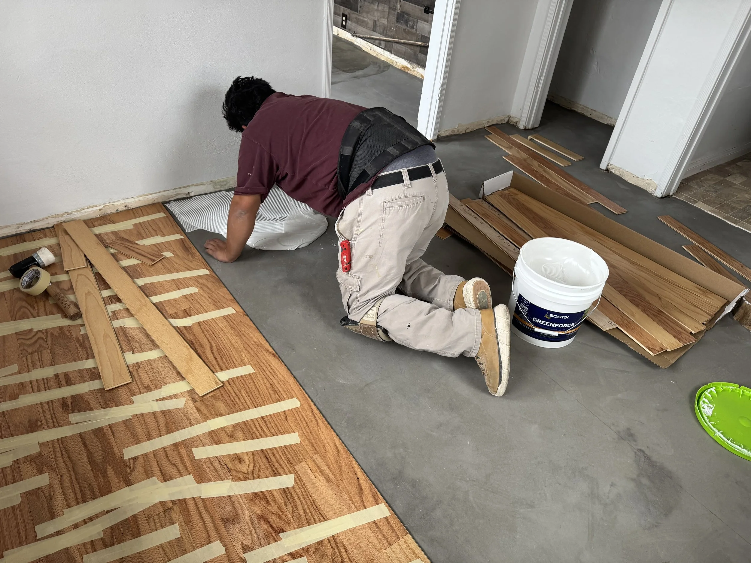 A person kneeling on the floor installing hardwood flooring, using masking tape to secure strips of wood to the subfloor. There is a bucket of adhesive and various wood planks around him.