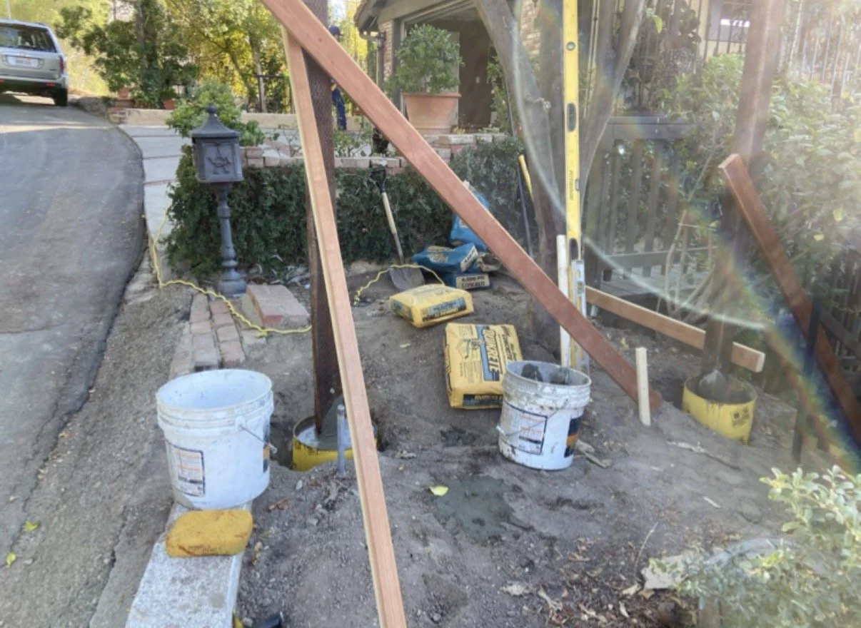 Construction site with wooden beams, buckets, and tools for building or repairing a small structure or fence near a sidewalk, with a parked car and a house with plants in the background.