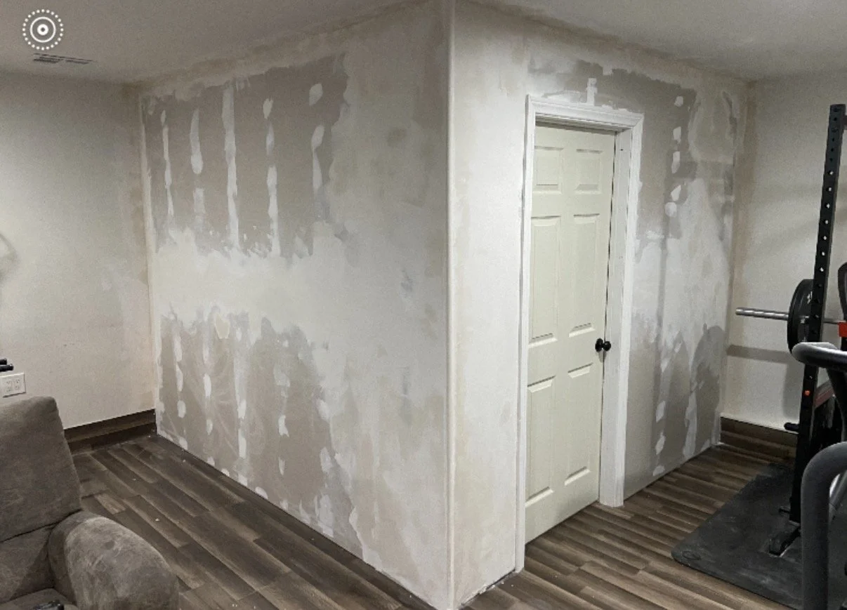 Interior wall under renovation with drywall mud patches, nearby door, and gym equipment in the corner.