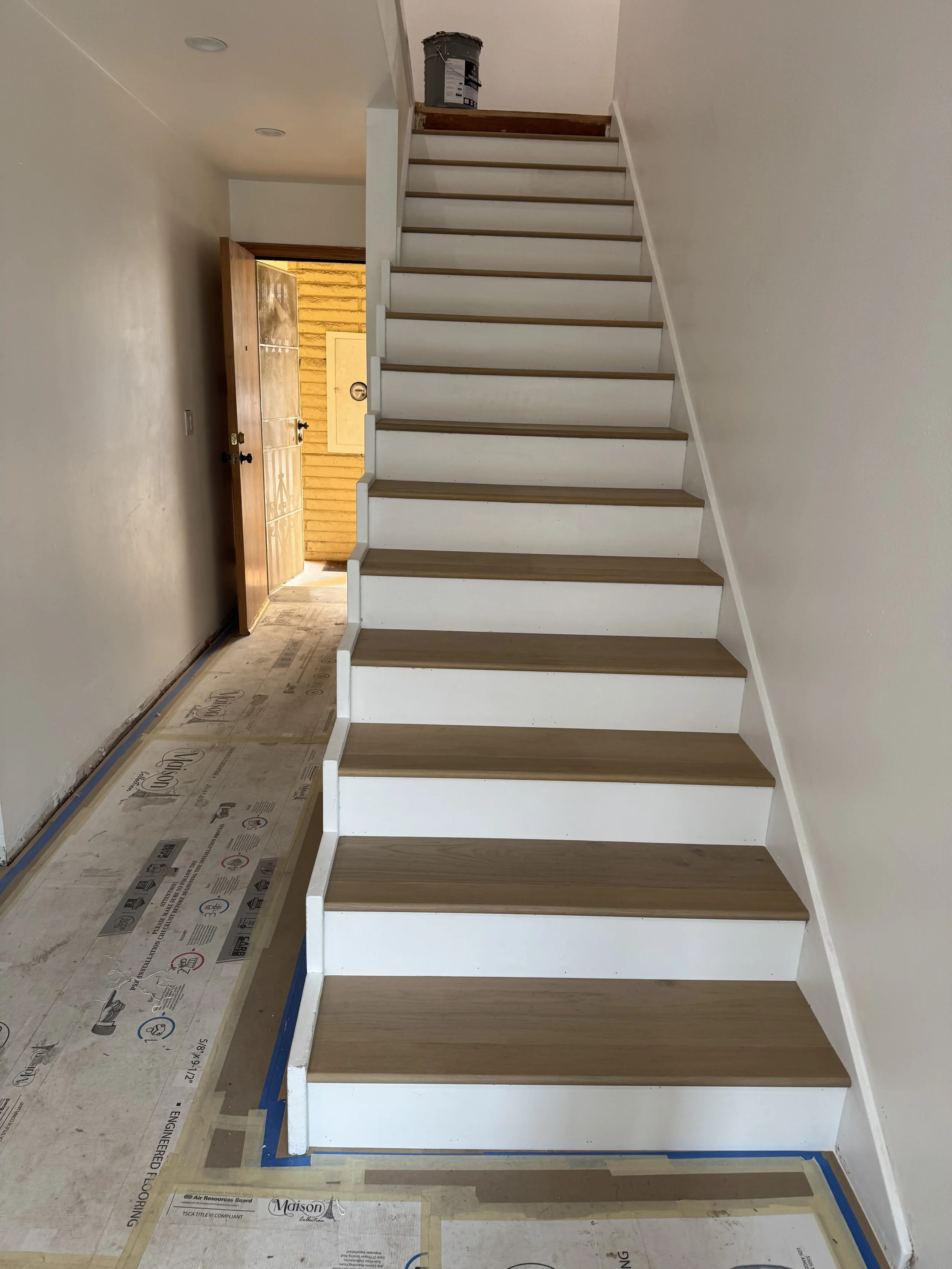 Indoor view of a staircase with brown wooden treads and white risers, leading upwards. The staircase is next to white walls, and at the top, there is a bucket. The bottom floor has clear plastic sheeting and painter's tape on the floor, indicating on