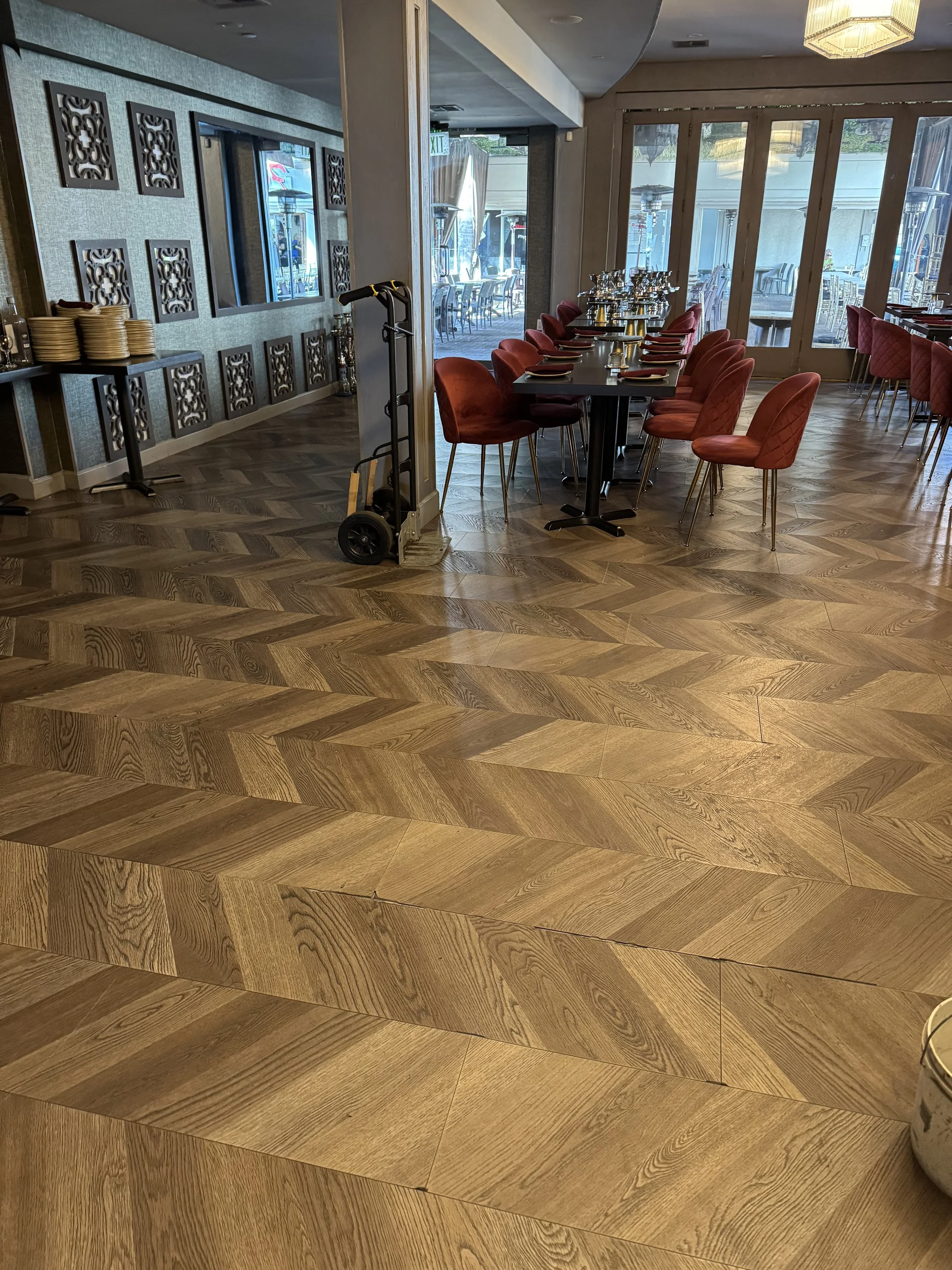 Empty restaurant with wooden flooring, red chairs around a black table, set for dining, and large windows showing outdoor seating.