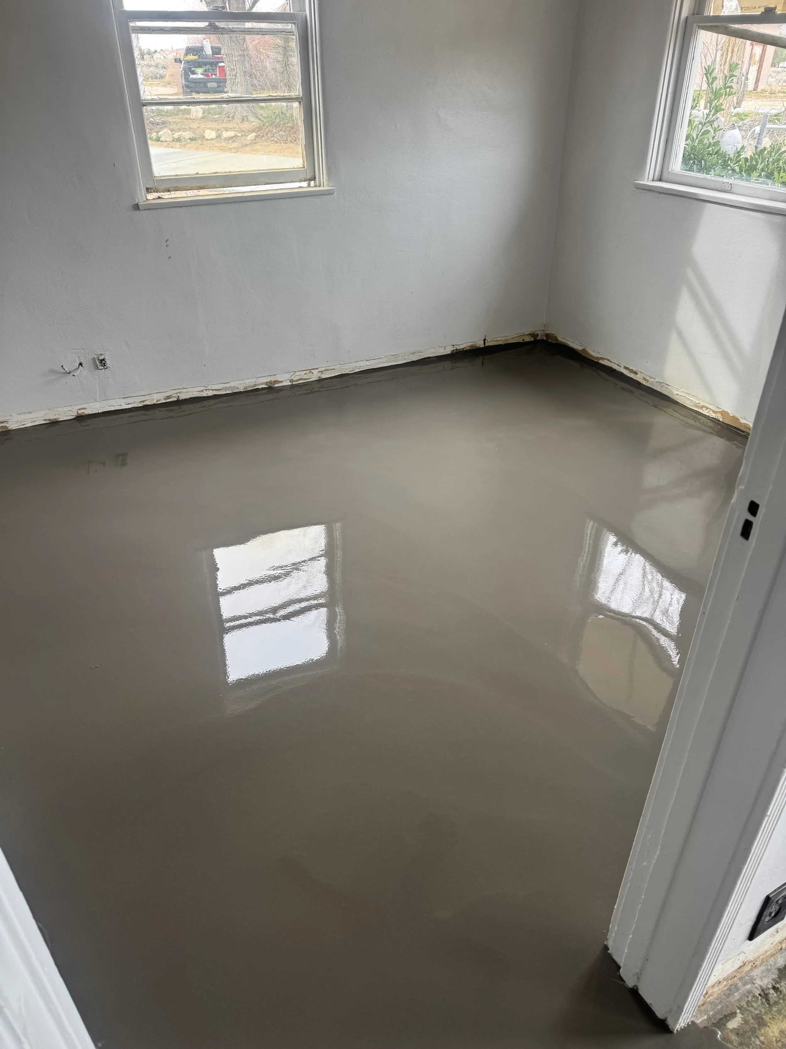 A room with newly poured epoxy concrete flooring that is glossy and smooth, with two windows letting in daylight, and walls painted white.