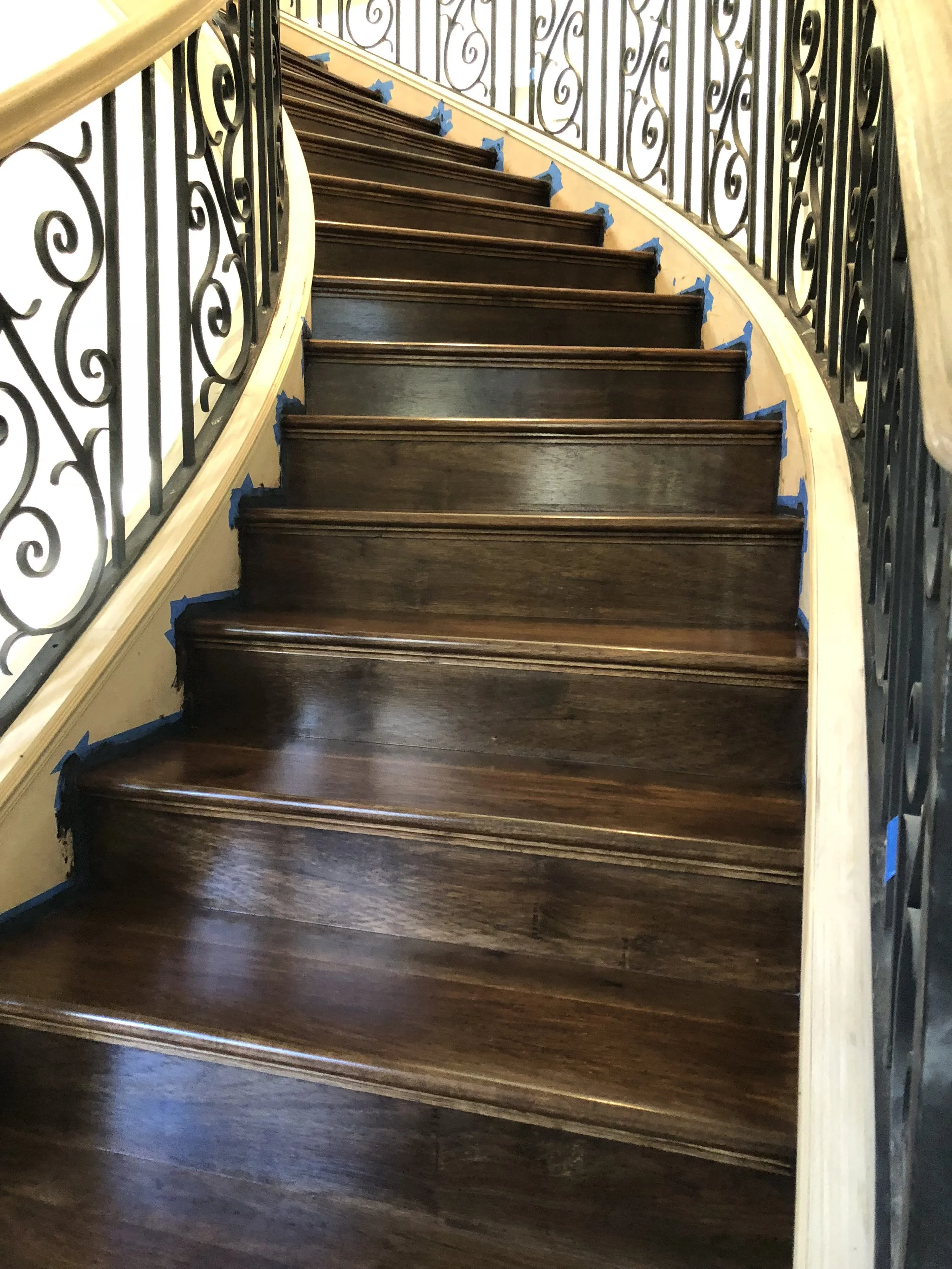 Photo of a spiral staircase with dark wooden steps and black wrought iron railings, with some blue painter's tape around the edges of the steps, suggesting ongoing maintenance or finishing work.