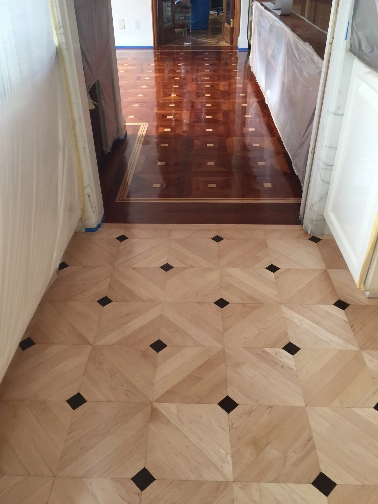 Interior view of a room with a transition from beige and black patterned tiles to polished dark wood flooring with inlaid geometric design.