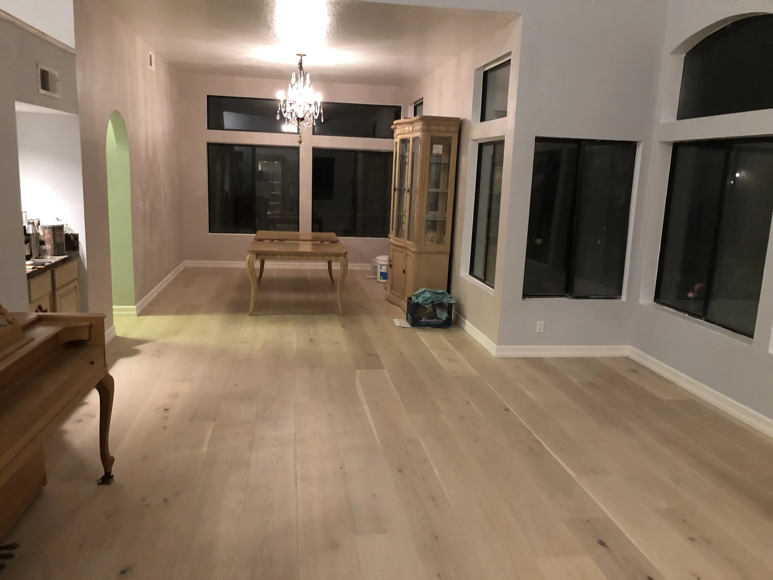 Empty living room with hardwood floors, large windows, chandelier, and a wooden table.
