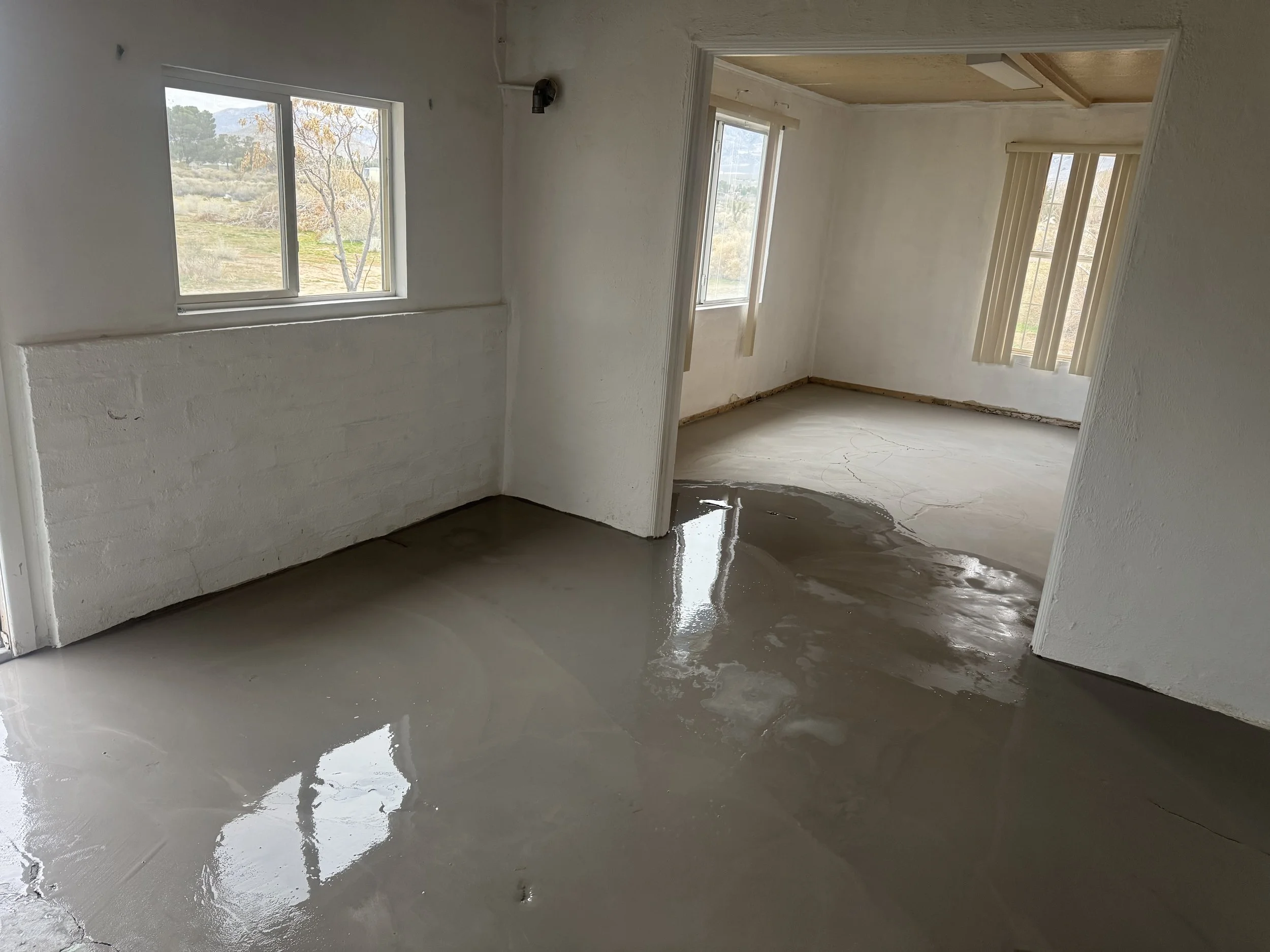 Empty room with wet concrete floor, white walls, and windows with blinds.