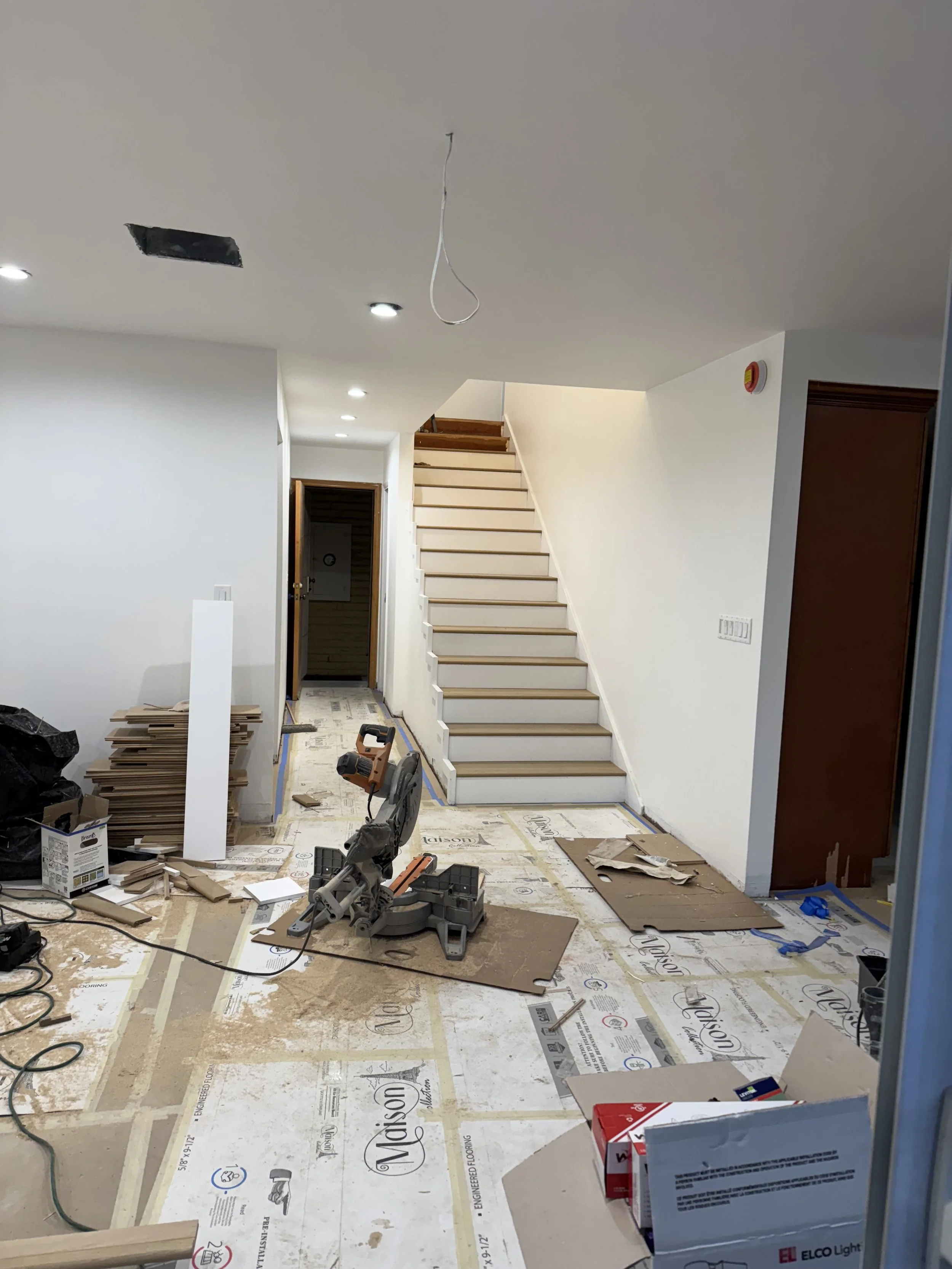 Interior view of a house under construction, showing a staircase, ongoing electrical work, construction tools, and building materials on the floor.
