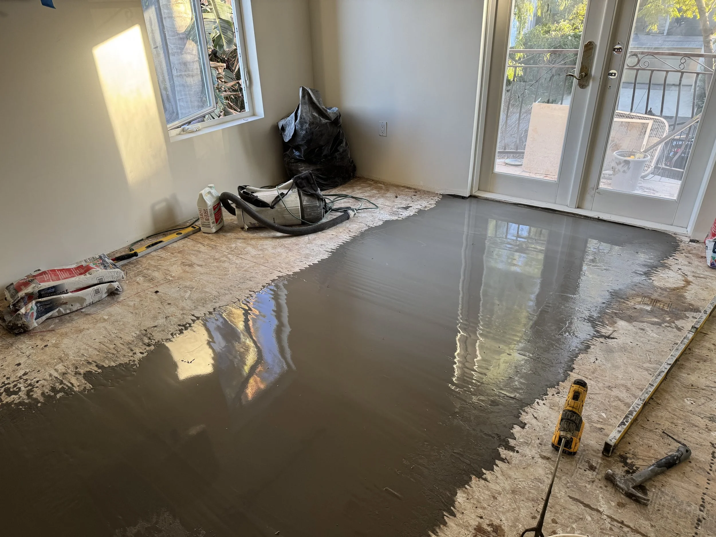 Room undergoing renovation with partially resurfaced floor; concrete sealant or epoxy coating being applied; tools and supplies like a level, drill, and cleaning chemicals are visible.