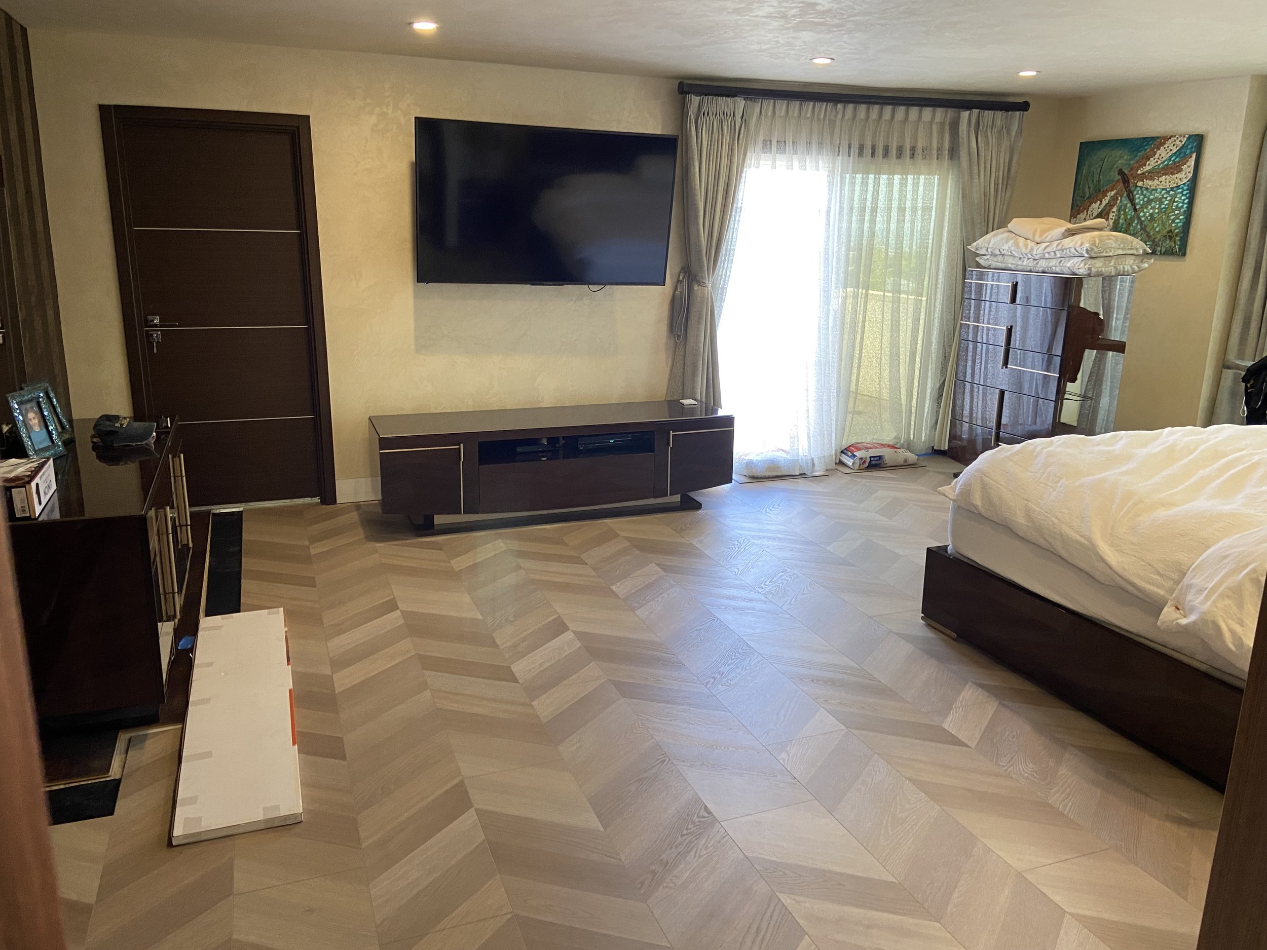 Living room with wooden flooring, a wall-mounted flat-screen TV, a closed wooden door, a dark wood TV stand, a sliding door with curtains leading outside, a dresser with pillows and cloth on top, a bed with white bedding, a small pile of items on the