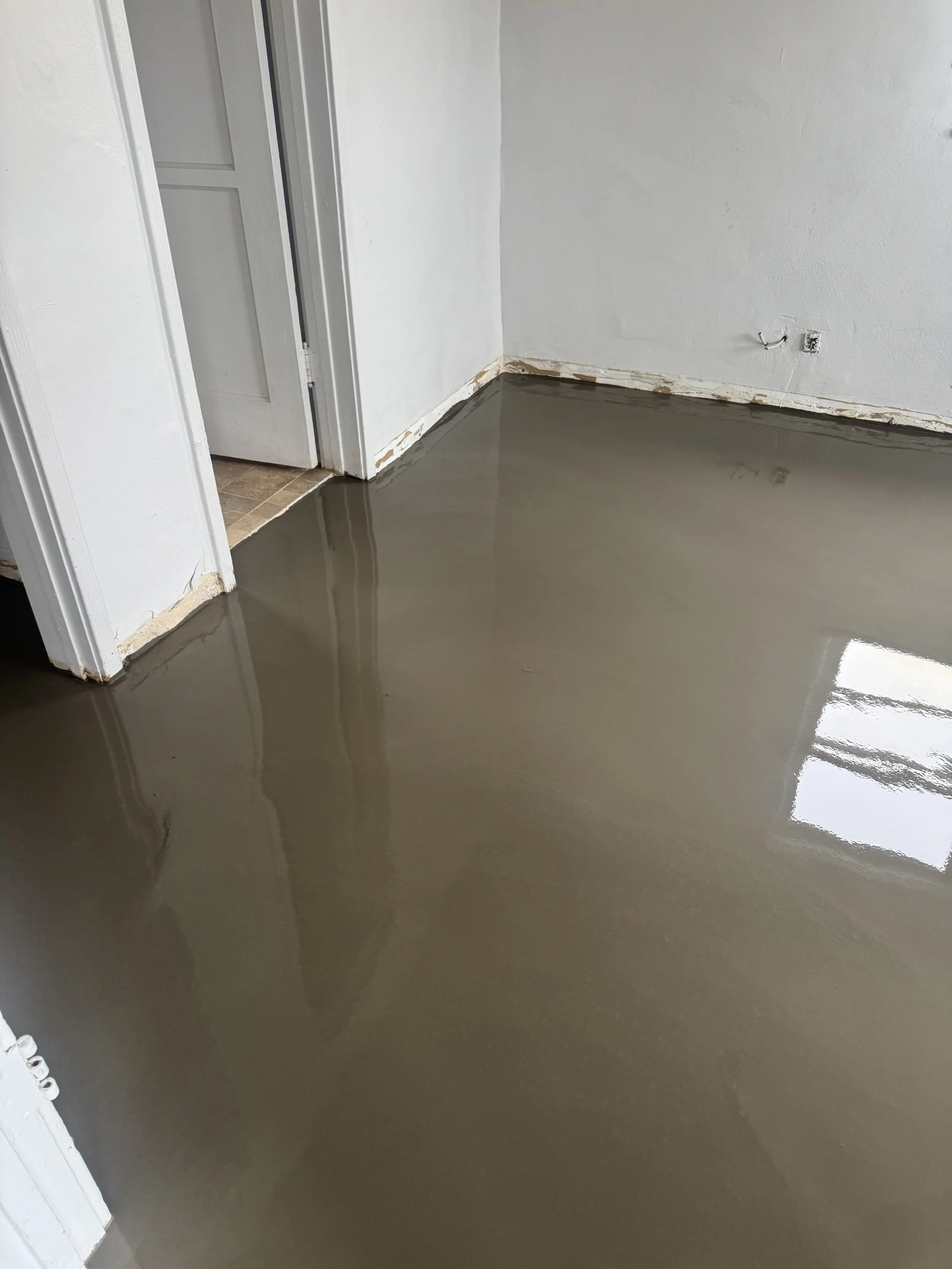 Interior room with freshly poured concrete floor, partially finished, with white walls and an open door showing part of another room.