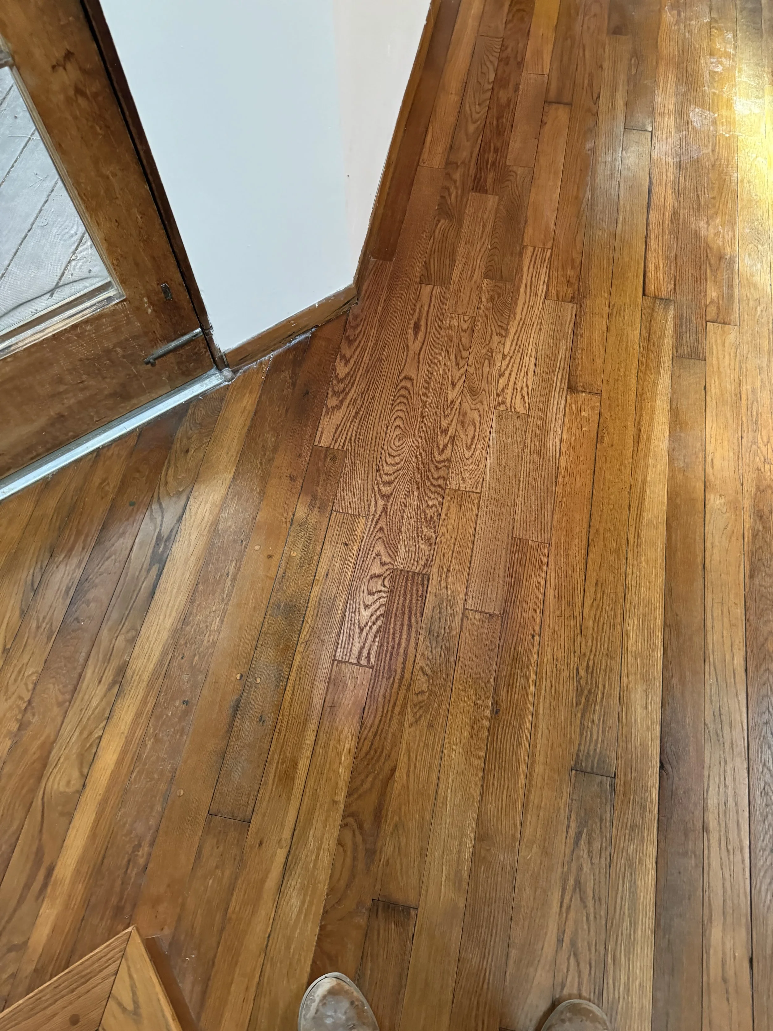 Close-up of a wooden floor with a partially old and worn finish near a doorframe, showing a section of hardwood flooring in a home interior.