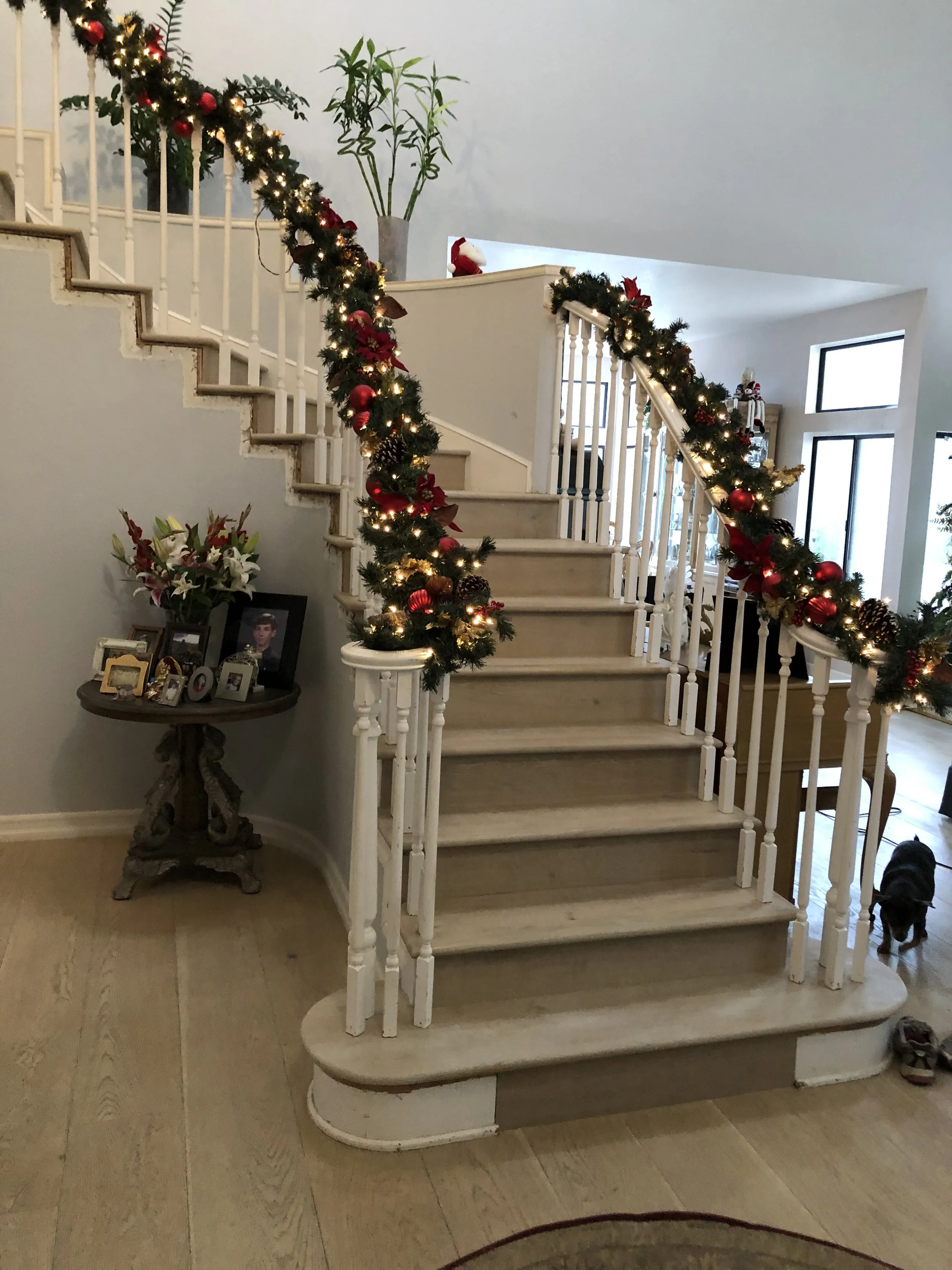Inside a house decorated for Christmas with a staircase adorned with garland and lights, a small table with photo frames and flowers, and a black dog near the bottom right corner.