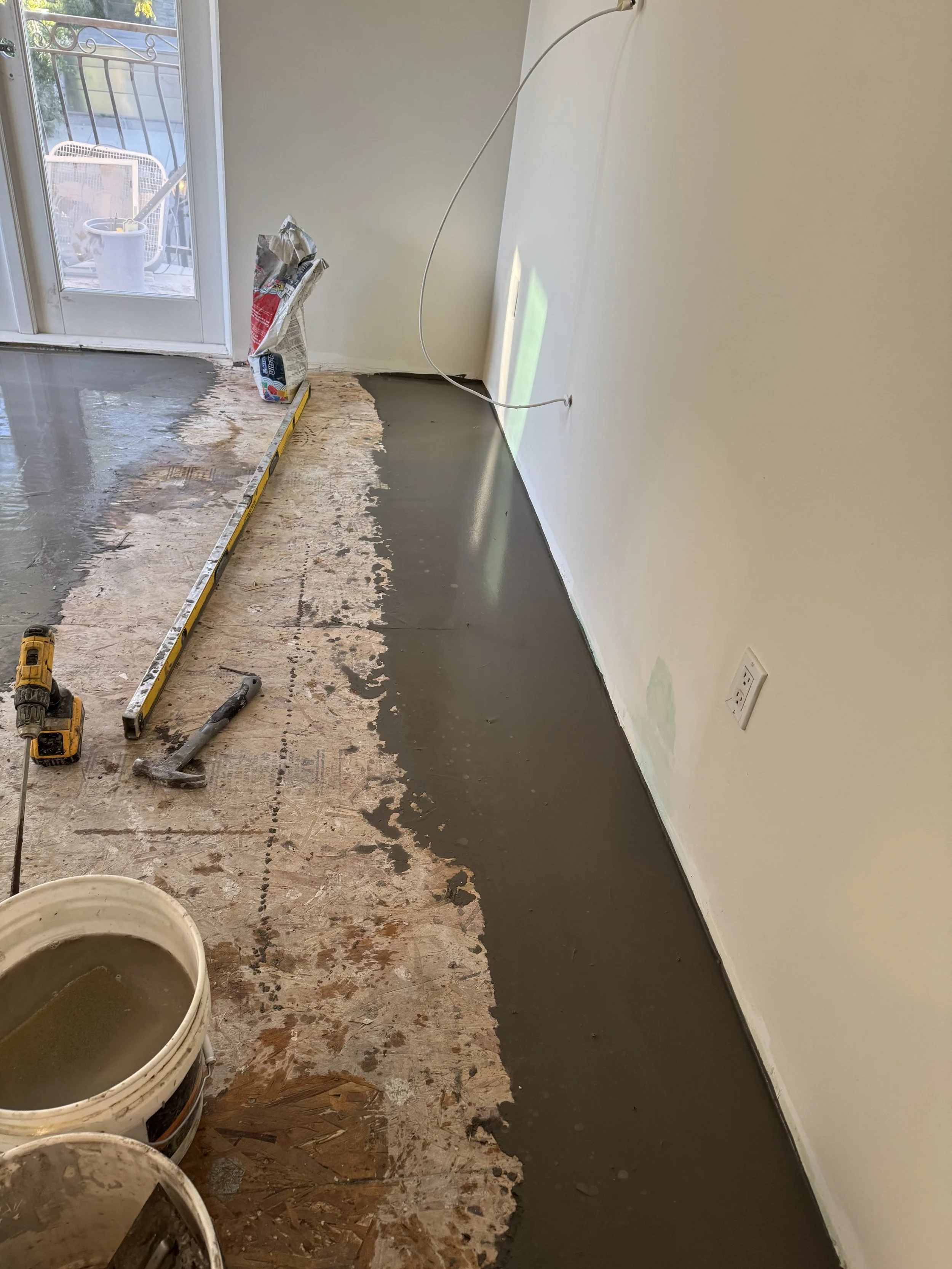 Interior room undergoing flooring renovation, with a fresh concrete layer being poured along the edge of the existing floor, construction tools, a level, and buckets visible.