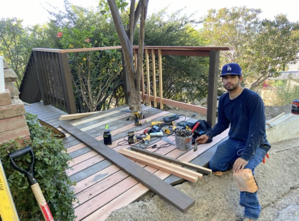 Man working on building a wooden deck, kneeling beside tools and wooden planks, wearing a blue Los Angeles Dodgers cap and dark shirt, with trees and shrubbery in the background.