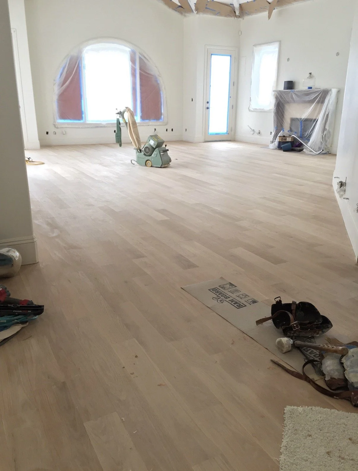 Room under renovation with light wood flooring, large arched window, glass door, and plastic coverings on other windows and fireplace.
