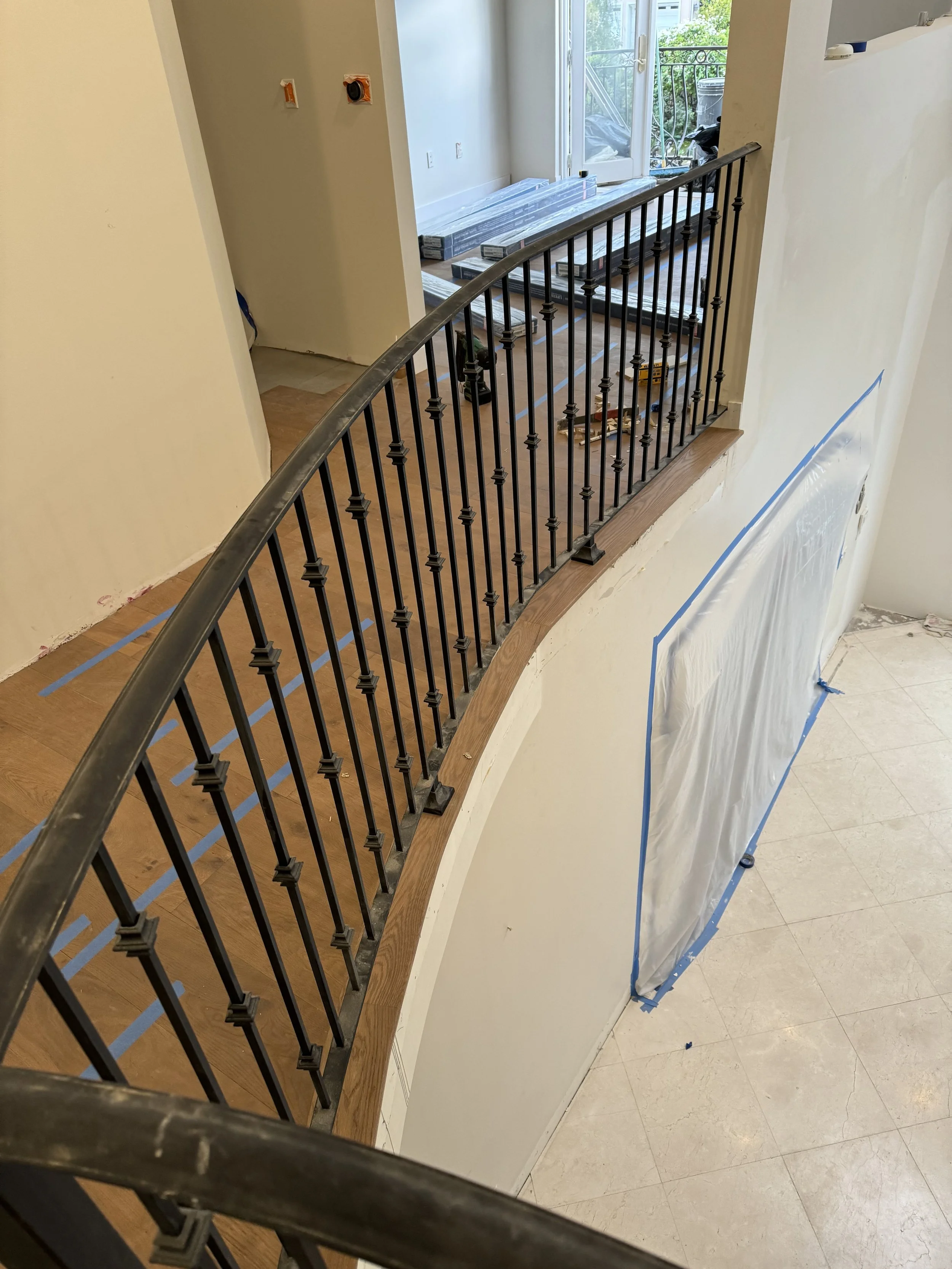 Partially finished interior staircase with black iron balustrade, wooden cap, and plastic covering on the wall below. There are tools and materials stacked on the floor in the background, and a balcony with a sliding glass door leads outside.