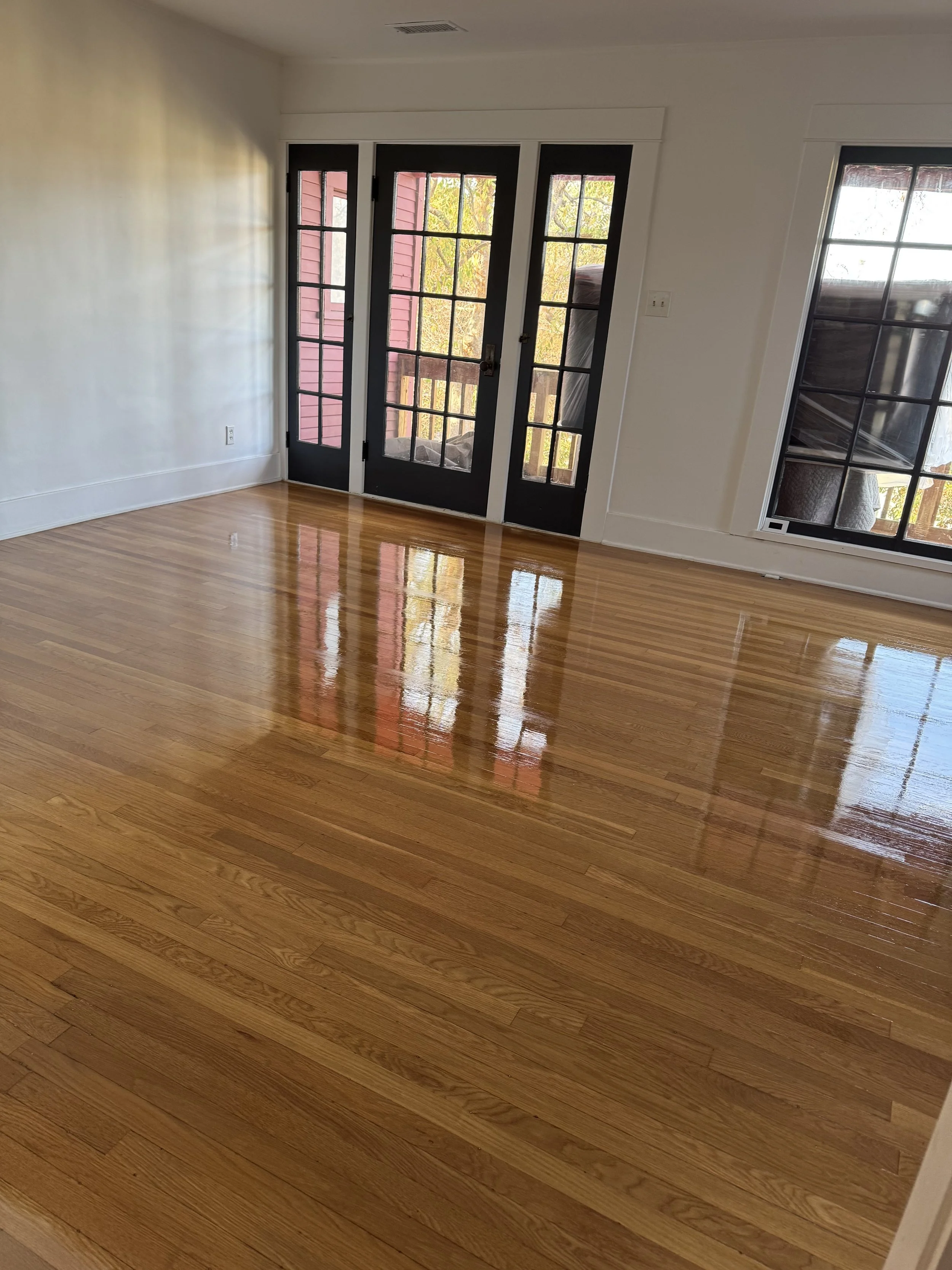 An empty room with polished hardwood floors, white walls, and black-framed glass doors leading to a balcony with a view of nature.