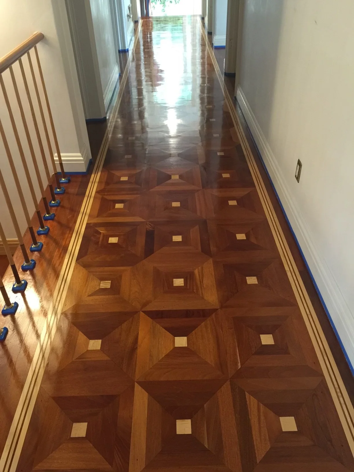 Polished wooden floor with a geometric pattern, bordered by a double line inlaid design, in a hallway with white walls, baseboards, and a railing on the left side.