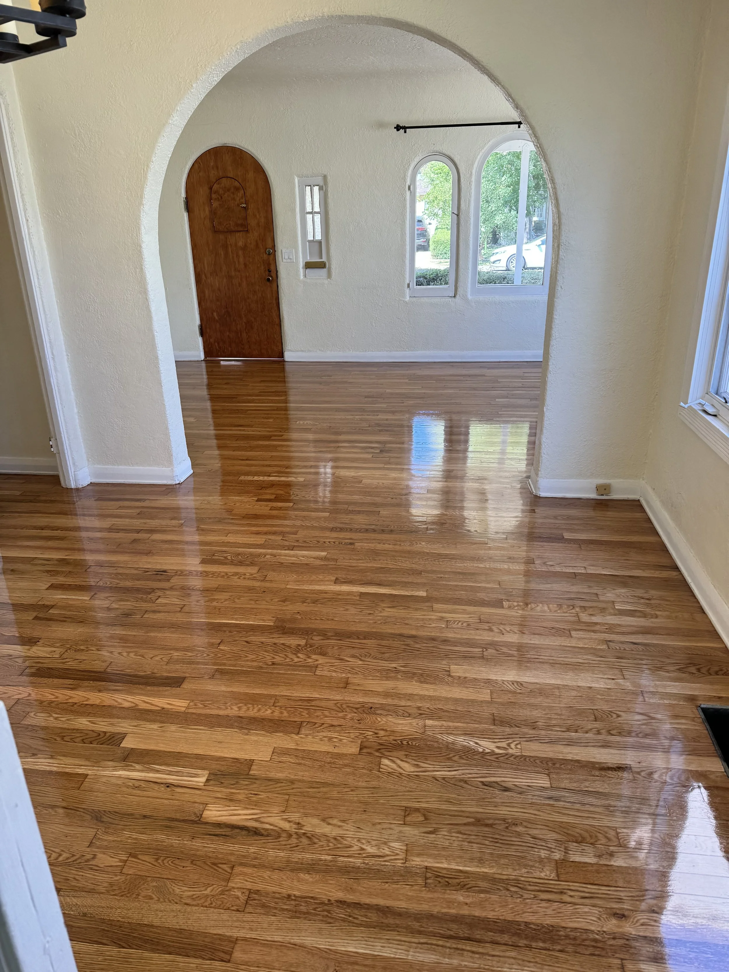 Empty room with shiny hardwood floors, light yellow walls, an arched doorway leading to another area, and windows with views of the outside greenery.
