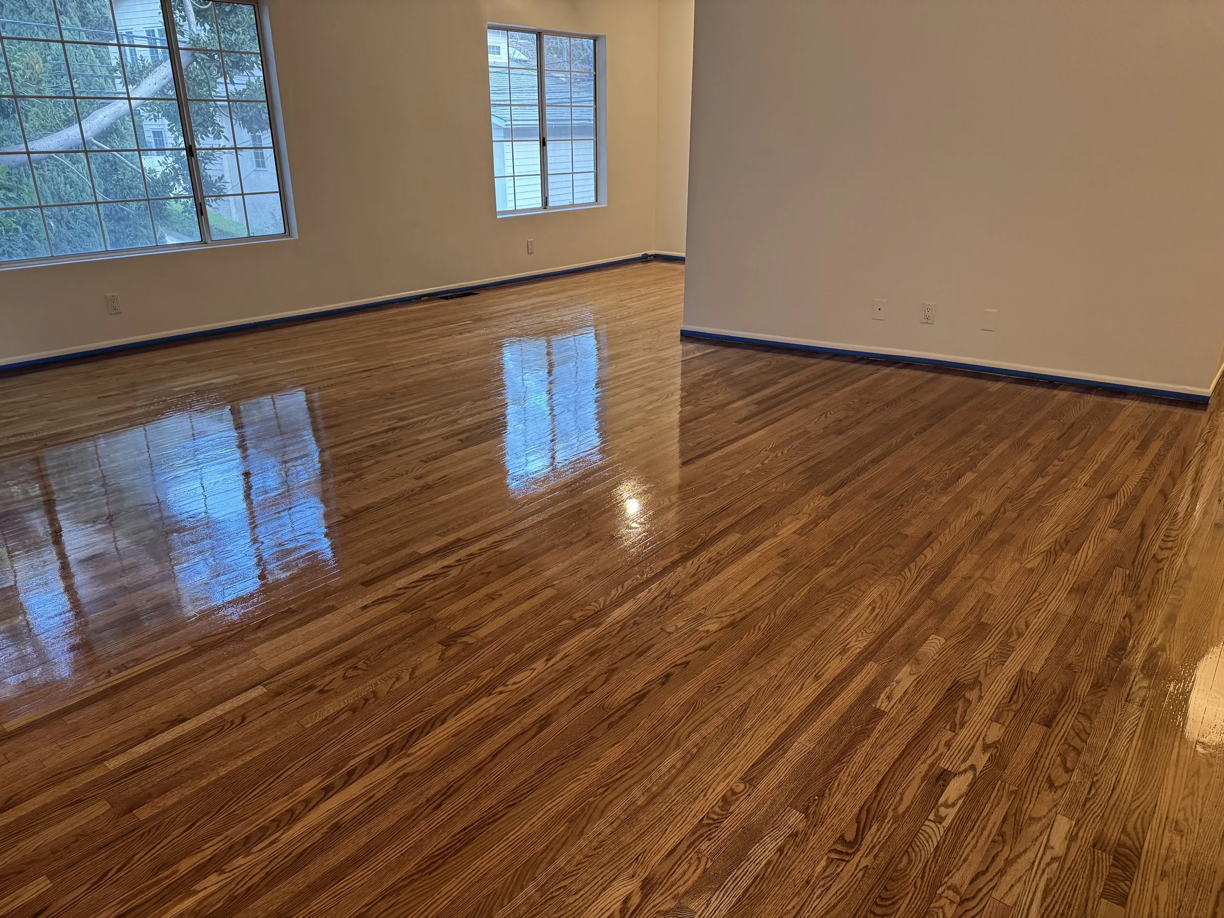Empty room with freshly polished hardwood floors and large windows that reflect the outdoors.