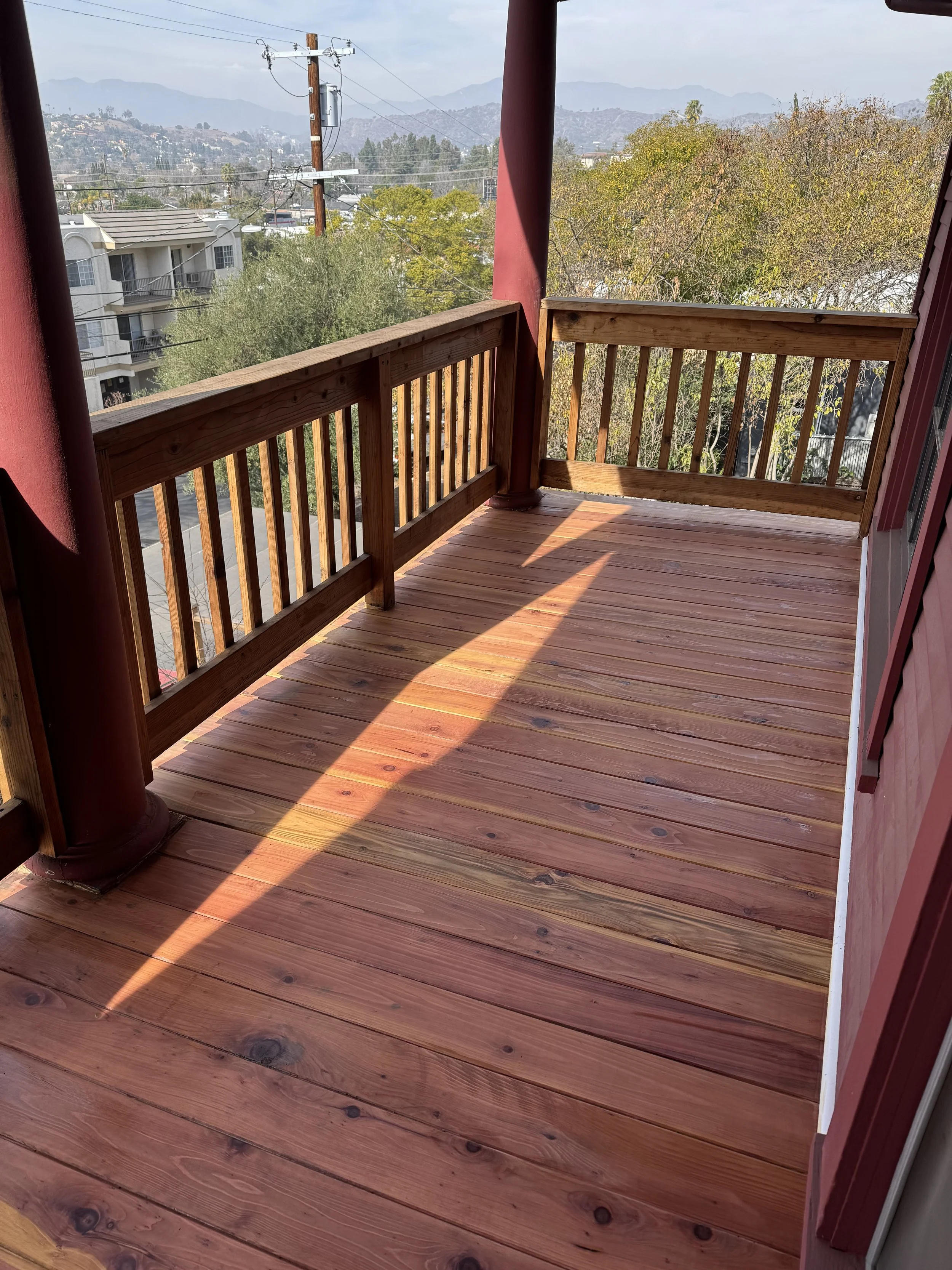 Empty wooden balcony with railing, view of trees, power lines, residential buildings, and distant mountains.