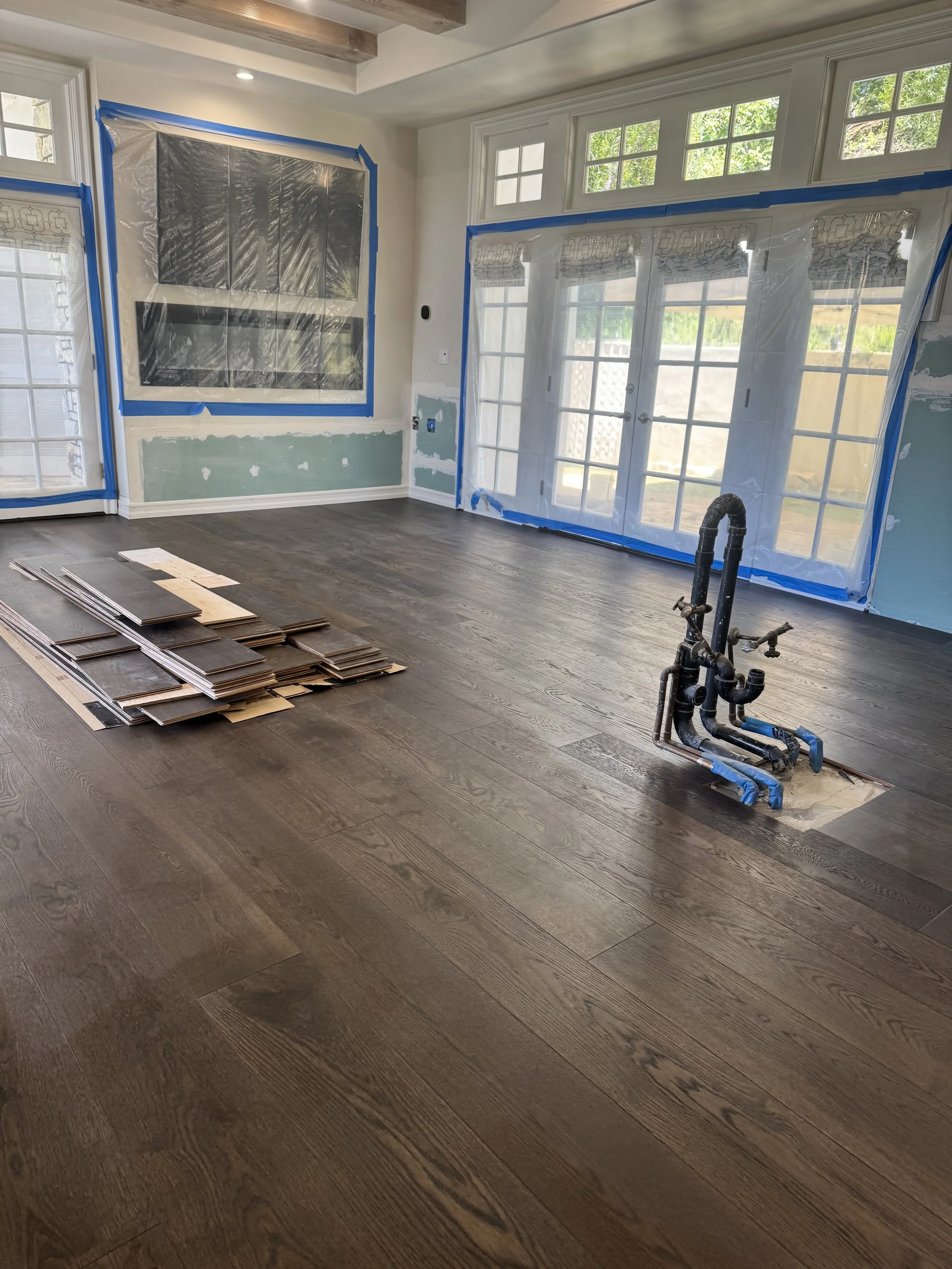 A room under renovation with dark hardwood flooring being installed. Piles of unused flooring planks are stacked on the floor. The room has large windows and sliding glass doors covered with protective plastic and blue painter's tape. There is an ope