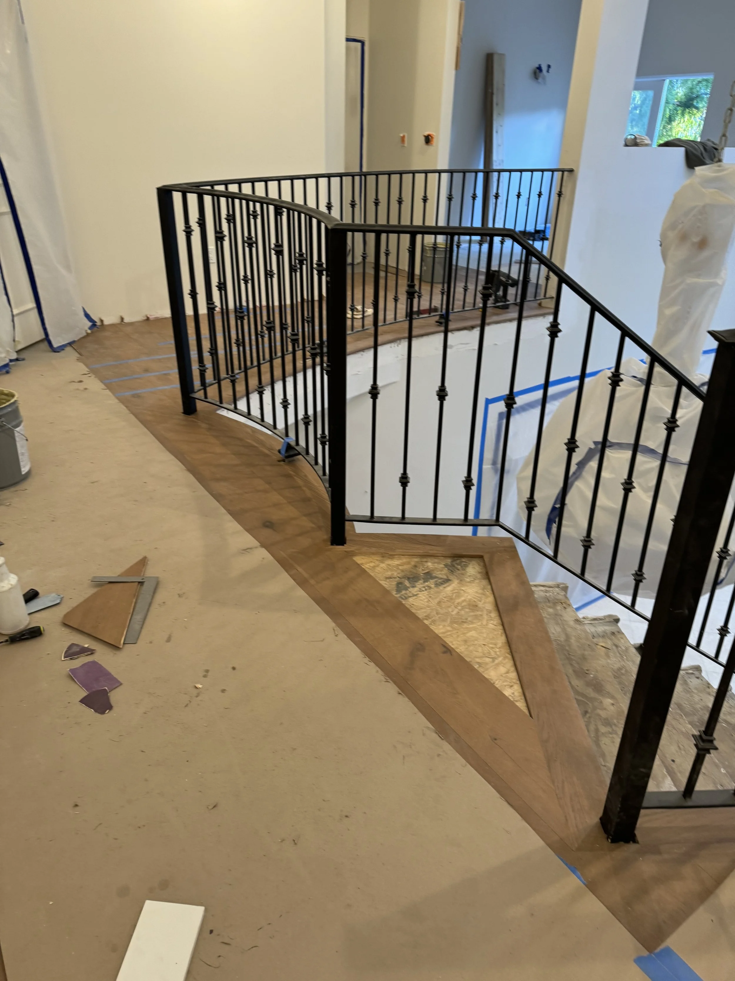 Interior of a house under renovation with a curved black metal railing along a staircase opening and partially installed hardwood flooring. Construction materials and tools are visible on the unfinished floor.